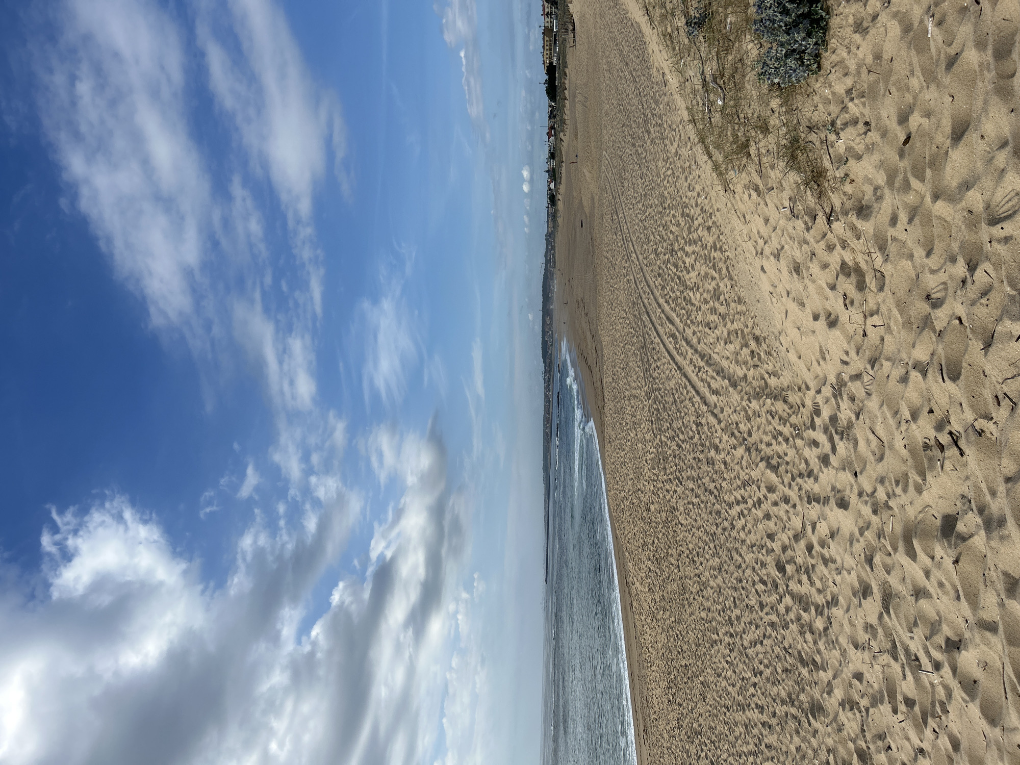 The image depicts a scenic beach on a bright, sunny day. The scene is dominated by the expanse of golden sand, which occupies the right side of the frame. The sand is textured with numerous footprints, suggesting recent human activity. Tire tracks are also visible, indicating the passage of vehicles. Further down the beach, a line of vegetation and some buildings can be seen in the distance, hinting at a developed coastline.
To the left of the sand is the calm, blue ocean, with small waves gently lapping at the shore. The water appears clear and inviting. The sky above is a vibrant blue, dotted with scattered white clouds. Some of these clouds are wispy and elongated, while others are more puffy and substantial. The lighting suggests it is daytime, likely late morning or early afternoon, given the bright sunshine and clear visibility.
The overall impression is one of a tranquil and pleasant day at the beach. There are no prominent objects or people clearly visible in the immediate foreground, but the footprints and tire tracks suggest a public beach that is frequented by visitors. The location appears to be a coastal area, possibly a popular tourist destination or a residential beach town. The weather is clearly favorable for outdoor activities.