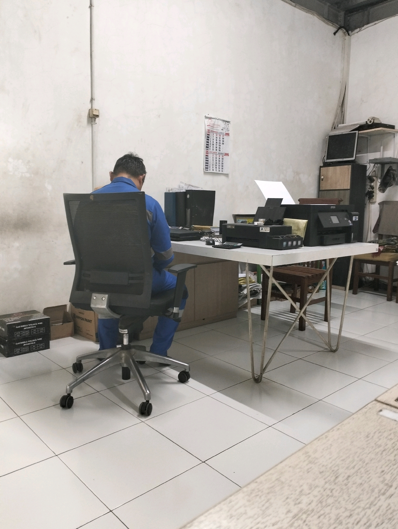 A man wearing a blue uniform is seated in a black mesh office chair, facing away from the camera, towards a white desk. The desk has several printers, a computer monitor, a calculator, and some papers on it. Behind the man, on the wall, is a calendar. The room appears to be an office or workshop, with tiled floors and white walls. The overall environment suggests a utilitarian workspace. This scene is located in the city of Gunung Sindur, Indonesia.