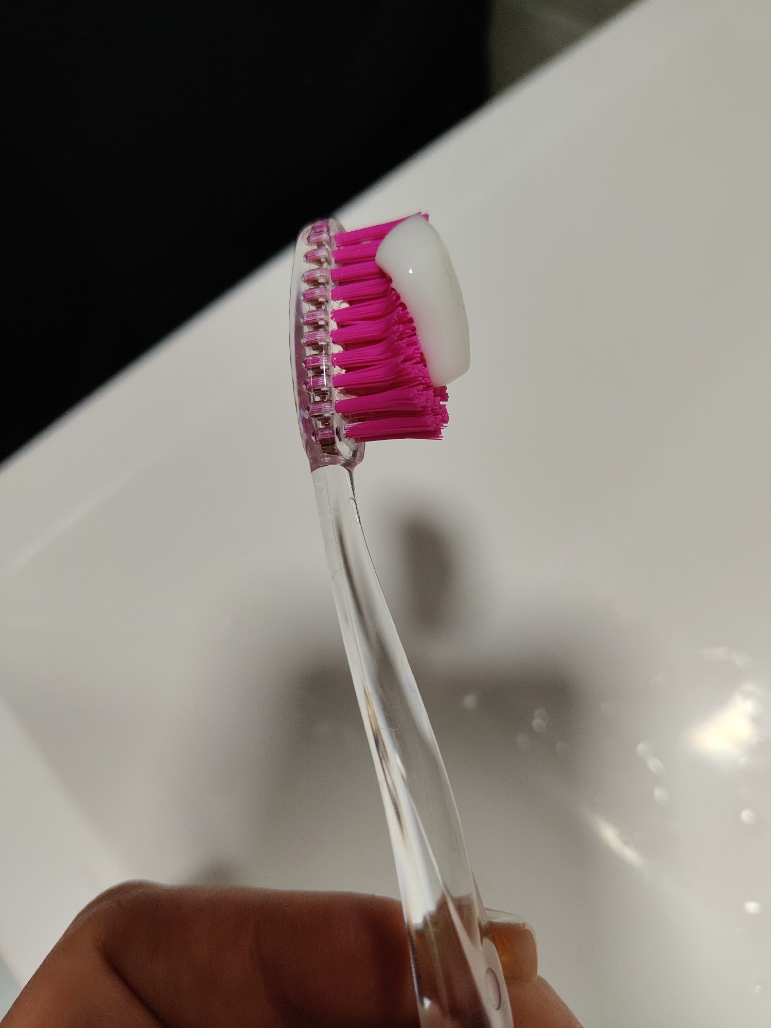 A close-up, indoor shot shows a pink toothbrush with white toothpaste on its bristles, held by a hand against a white sink background. A dark area is visible behind the sink.