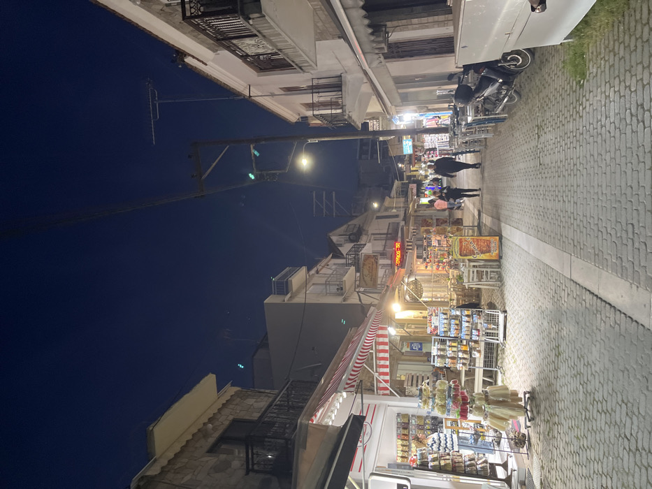 The image depicts a narrow cobblestone street in Limenaria, Greece (latitude: 40.627, longitude: 24.575) at night. The street is lined with shops and restaurants on both sides, with red and white striped awnings over some of the storefronts. There are a few people walking down the street, all facing the same direction. The sky is dark blue and there are several streetlights illuminating the street. The street is relatively empty with a few scattered tables and chairs in front of some of the shops. A motorcycle is parked on the right side of the street. The scene is dimly lit and appears to be relatively peaceful.