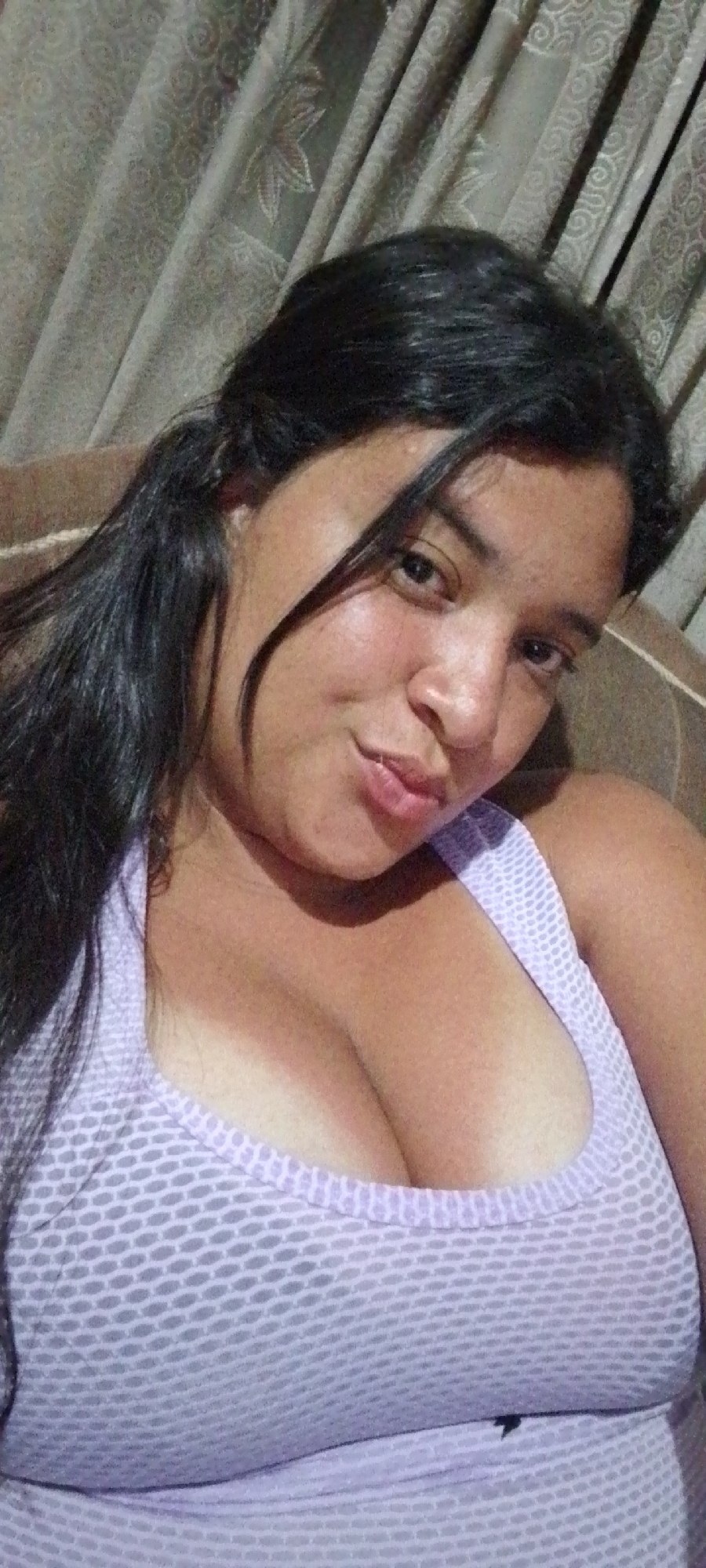 A woman with dark hair is posing for a selfie, wearing a light purple patterned tank top. She is looking directly at the camera with a slight smile and her hair is pulled back. The background appears to be a room interior with light-colored curtains. The specific location in La Cabrera, Venezuela cannot be determined from the image.
