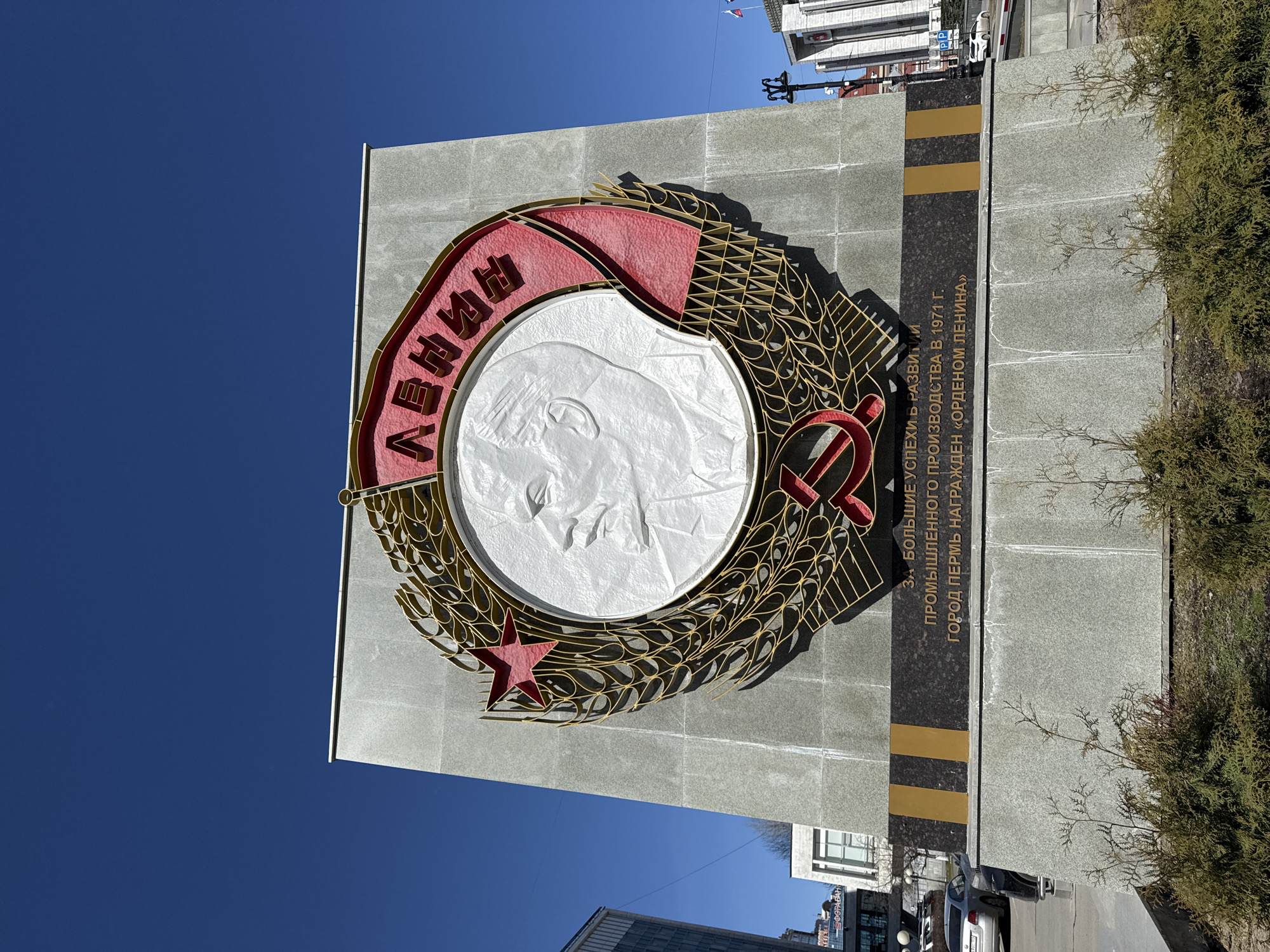 The image features a large stone monument in Perm, Russia, dedicated to Vladimir Lenin. The monument is gray with a large circular emblem prominently displayed. Inside the emblem is a raised white relief profile of Lenin's face. The emblem is surrounded by a golden wreath and a red banner with the name "Ленин" (Lenin) in Russian. A red star and hammer and sickle symbol are also part of the emblem.
To the right of the emblem is a black plaque with gold stripes, inscribed with Russian text stating that the city of Perm was awarded the Order of Lenin in 1971 for significant achievements in industrial development. The monument is located outdoors, with blue sky visible in the background. Trees and buildings, including cars and commercial establishments, are visible at the base of the monument, indicating that it is situated in an urban setting, likely near a street. Flags are seen on top of a building in the background.
