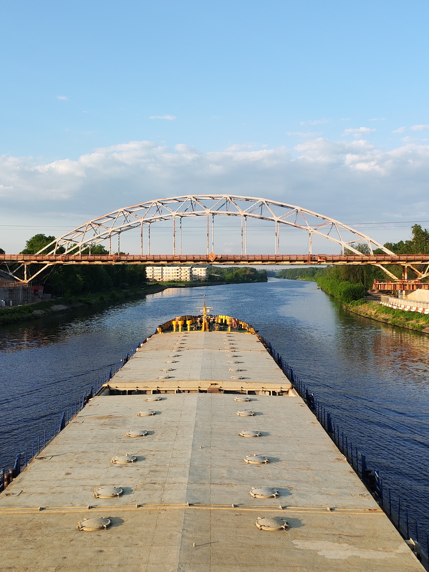 The image shows a large, flat-top barge moving along the Moscow Canal near Dmitrov, Russia. The barge's deck is concrete-colored and features a repeating pattern of circular access hatches. The canal's water is dark blue, and ripples indicate movement. The banks of the canal are lined with trees and some man-made structures, including a building that appears to be a multi-story apartment complex. Above the barge and spanning the canal is a rust-colored arched bridge with a white metal framework. The sky is blue with some scattered clouds. There are no people visible in the image.