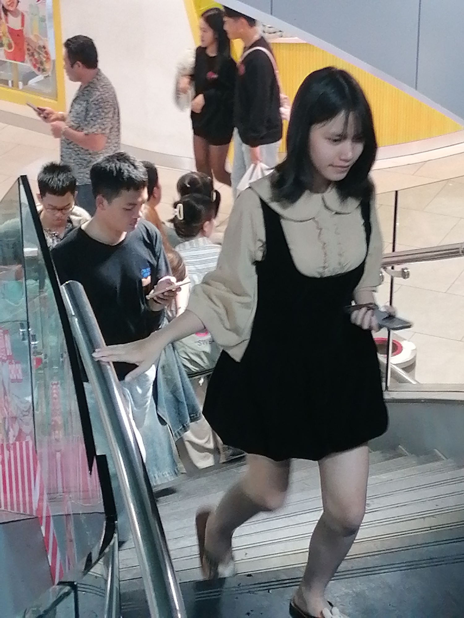 The image depicts a group of people on a staircase, likely within a shopping mall or public building in Da Lat, Vietnam. A young woman in the foreground is wearing a black dress over a light-colored blouse with a Peter Pan collar. She is holding a smartphone in her right hand. Behind her and to her left, a man in a black t-shirt is looking at his phone. Further back, several other individuals are visible, including a man in a patterned t-shirt holding a phone, and two women, one in a black top and the other in a striped shirt. The staircase appears to be made of metal and concrete. The background includes signage and yellow architectural elements, common in commercial spaces. The lighting suggests an indoor environment.