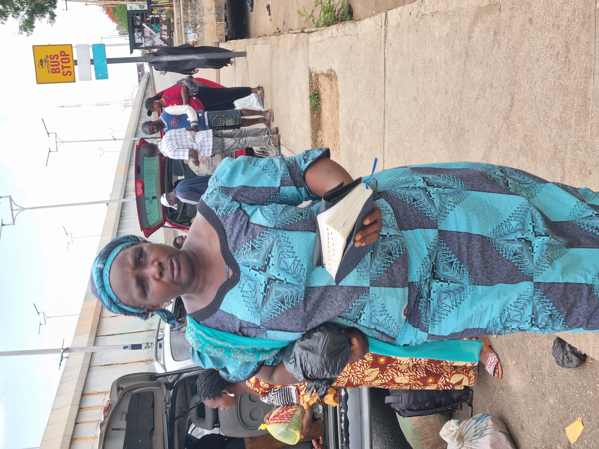 A woman holding a book stands on a sidewalk. She is wearing a blue and gray patterned dress and a matching headwrap. People and luggage are behind her, positioned near a red car with an open trunk. A bus stop sign is visible in the background. The location appears to be near an overpass in the city of Jos, Nigeria.