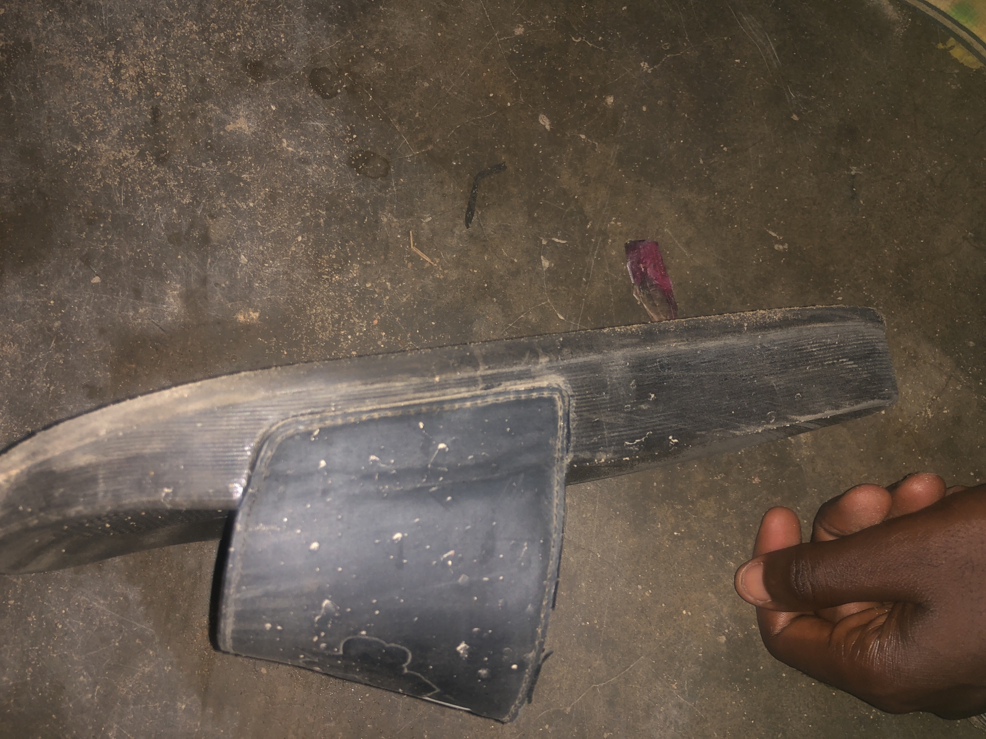 A dark-colored sandal is placed on a dirt ground. A hand is visible on the left side of the image, holding the sandal. The sandal is worn and appears to be made of rubber. There are some white specks on the sandal and the ground, which could be dust or debris. The ground appears to be a mixture of dirt and concrete. There is also a small piece of purple material to the right of the sandal. The image is taken from a top-down perspective.
