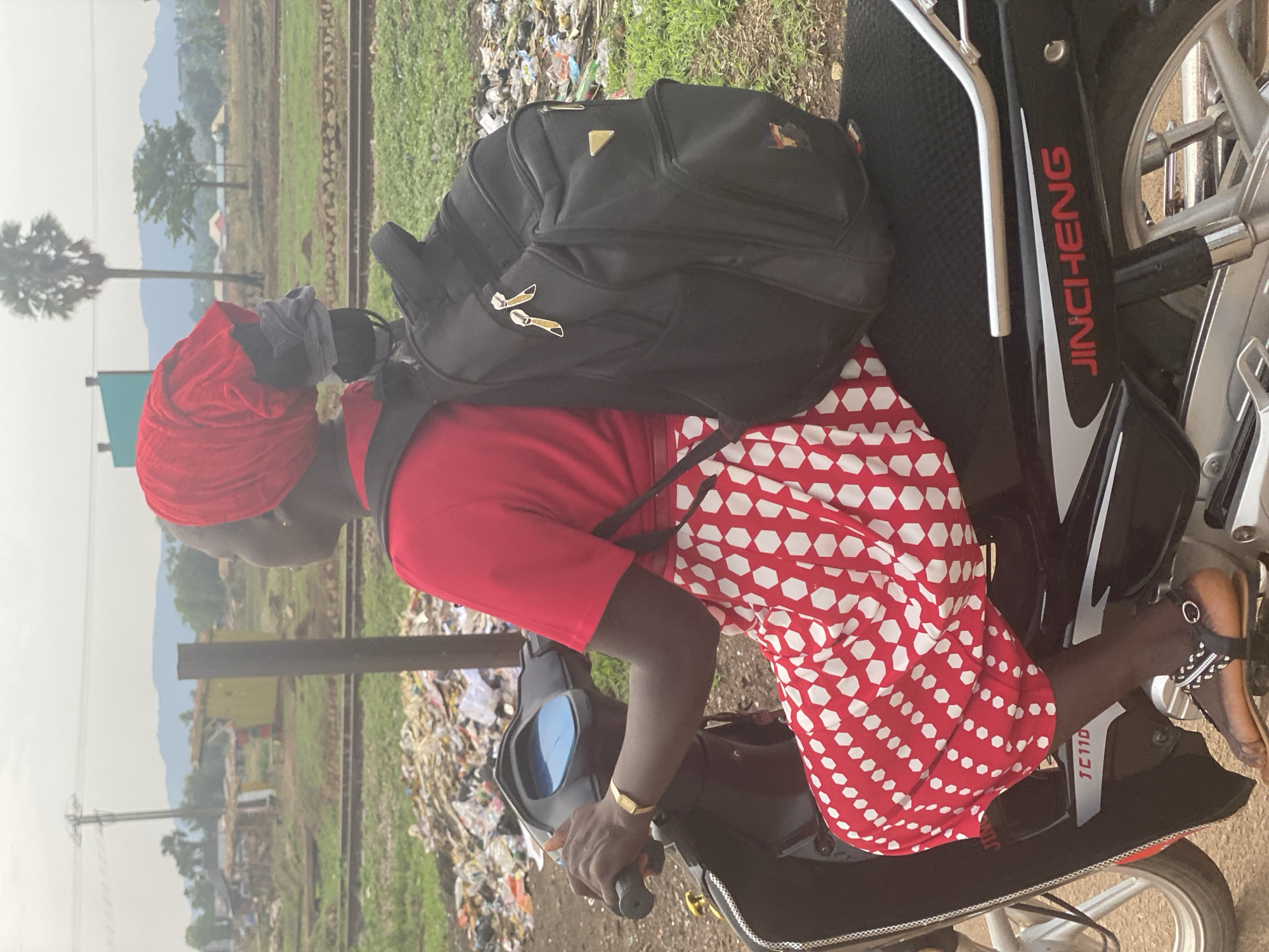 A person is seated on a black motorcycle, facing towards the left of the frame. She wears a vibrant red headwrap, a red short-sleeved top, and a long red skirt with a white hexagonal pattern. A black backpack with gold zipper pullers is strapped to her back. Her right hand rests on the motorcycle's handlebar, revealing a gold-colored bracelet on her wrist. The side of the motorcycle clearly displays the brand "JINCHENG" in red lettering and "TC110" in white.
The background reveals railway tracks running horizontally behind the motorcycle. Beyond the tracks, there is green vegetation, scattered simple structures, and hazy distant mountains or hills under an overcast or diffused sky. A tall palm tree and a utility pole are visible further in the distance. To the right of the railway tracks (and behind the motorcycle), a significant pile of trash and debris indicates a heavily littered area. The scene takes place during the daytime, likely in a rural or semi-urban setting, consistent with the provided context of Maigizo, Nigeria.