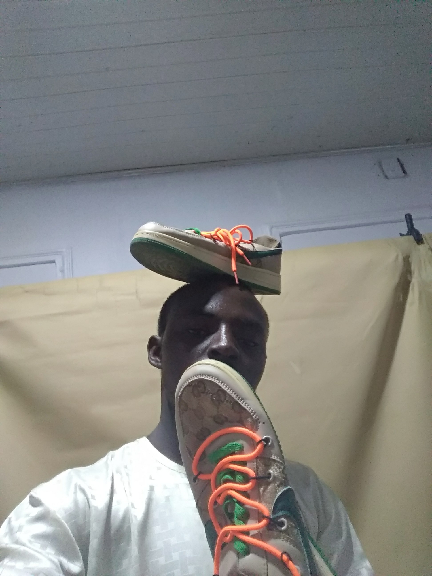A man is holding up two sneakers. One sneaker is on his head and one is in front of his mouth. The laces of the sneakers are orange. The background is a yellowish-brown curtain and a light gray ceiling. The location is Unknown, Unknown.