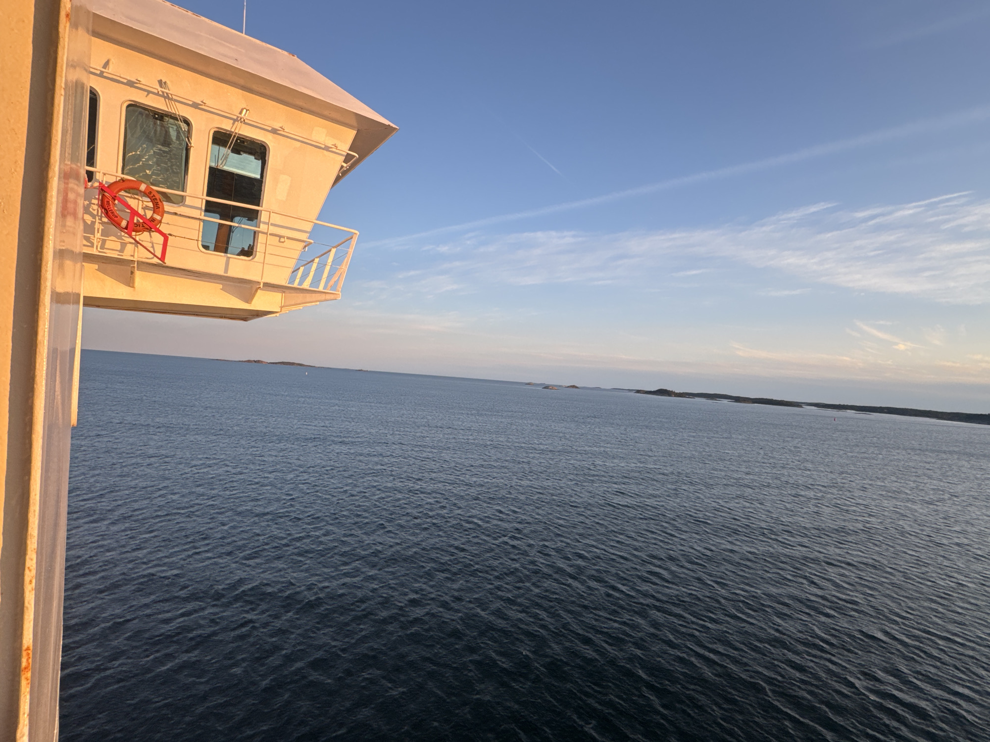 The image shows a view from a ship at sea, likely near Nynäshamn, Sweden. In the foreground, a portion of the ship's bridge or a similar structure is visible, painted yellow-orange and illuminated by sunlight. A red lifebuoy with the inscription "STERK" is hanging on the exterior wall of this structure. The sea stretches out to the horizon, appearing dark blue with small waves. In the distance, islands or a coastline are visible. The sky is a clear blue with some wispy white clouds and a faint contrail. The overall impression is one of a calm day at sea, possibly during sunrise or sunset given the warm lighting on the ship's structure.
