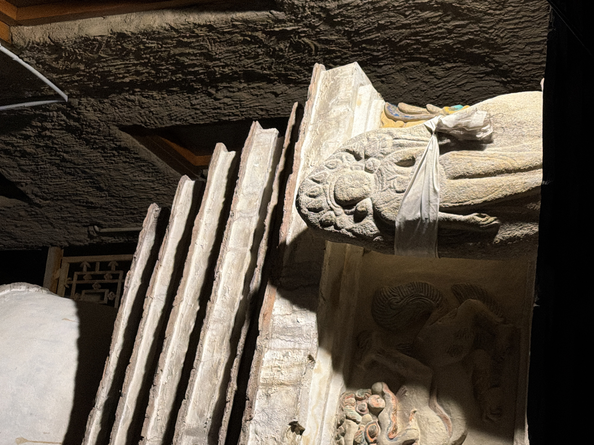 The image shows a close-up of an ancient stupa, a Buddhist reliquary monument, at night in Leh, India. The stupa features tiered levels with carved stone details, including figures and decorative elements. A prominent stone carving of a human-like figure, possibly a deity or protector, is visible in the foreground, wrapped with a white cloth. To the left, another carved element depicts a mythical creature, likely a dragon or lion. The background consists of a textured, dark wall, possibly rock or ancient plaster, illuminated by artificial light that casts dramatic shadows on the stupa. The overall scene evokes a sense of antiquity and spiritual significance.
