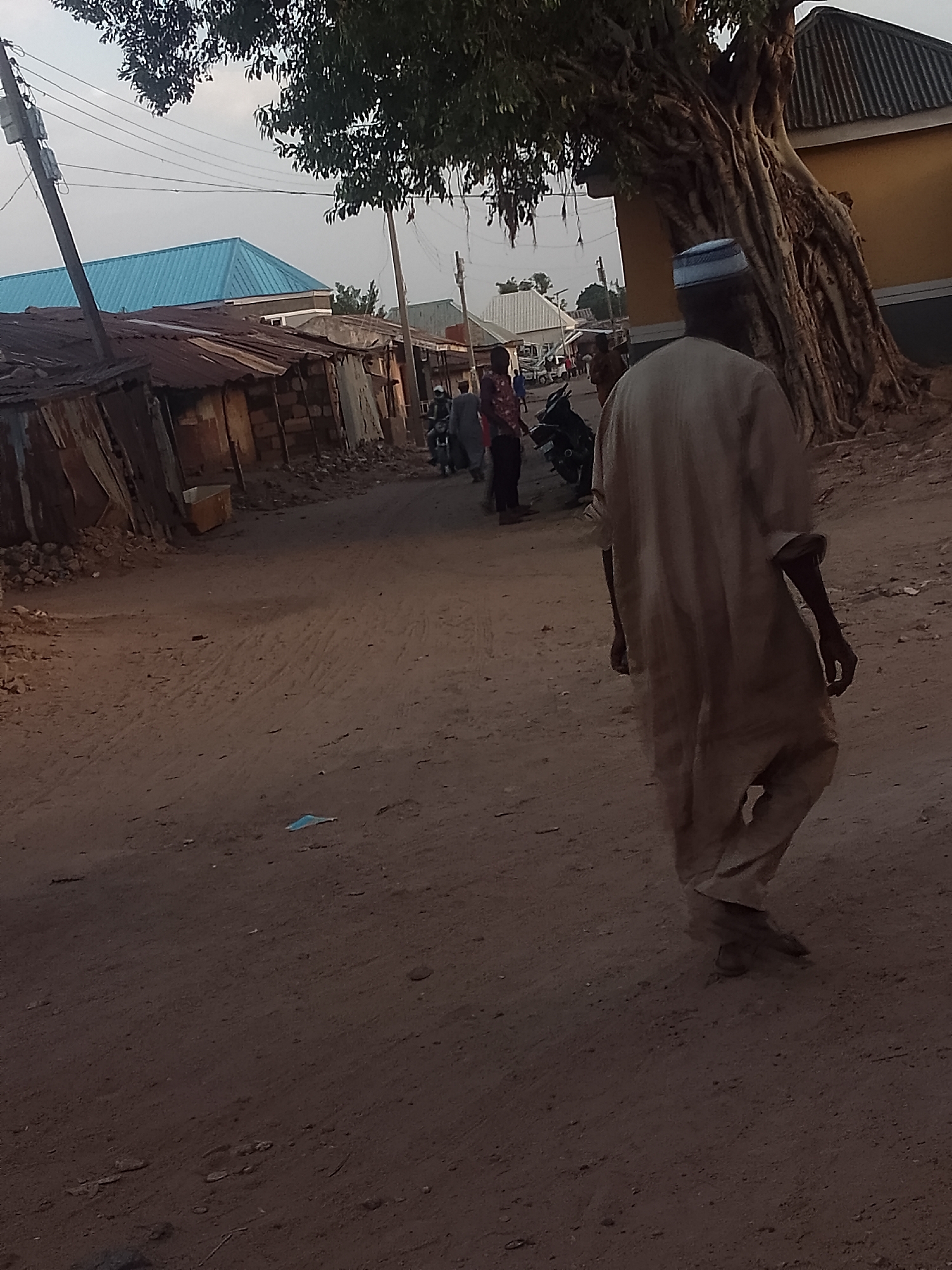 The image depicts a dirt road in Pantame, Nigeria. A man wearing traditional attire walks away from the camera. Several other individuals and motorbikes are further down the road, between small, corrugated metal structures. A large tree with visible roots stands beside a building. Power lines are visible above the structures.