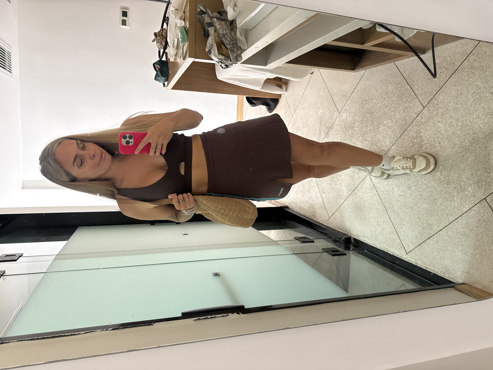 A woman with blonde hair is taking a mirror selfie in a hotel room. She is wearing a brown sports bra and a brown skirt. She is holding a straw bag in her left hand and a pink iPhone in her right hand. The room has tiled floors and glass doors. There is a wooden vanity with various items on it, including a green brush and a small leopard-print stuffed animal. The background suggests the location is Pudong, China, likely a hotel, given the mirrored closet and vanity area.
