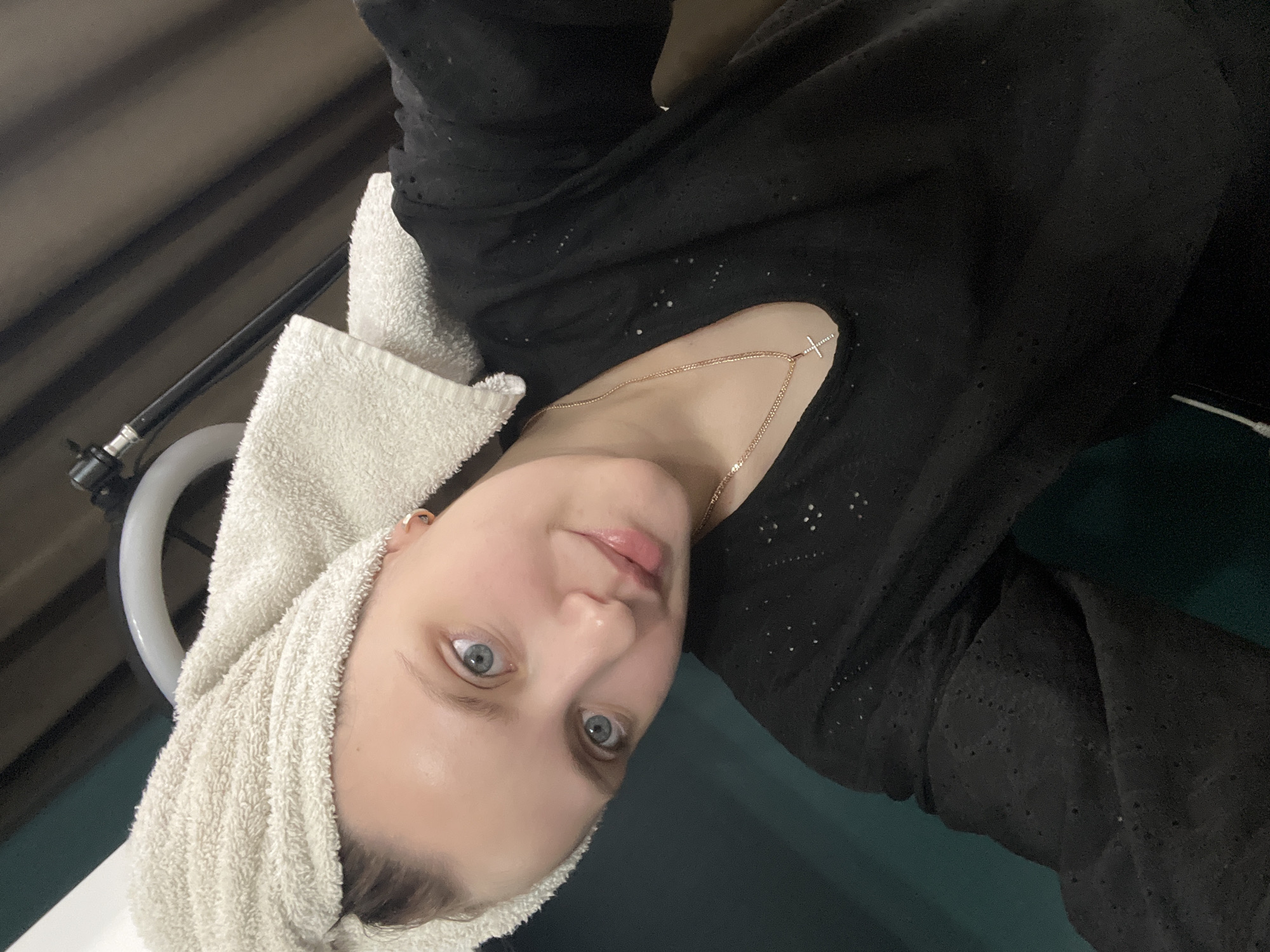 The image presents a close-up, head-and-shoulders portrait of a young woman. She is looking directly at the camera with wide, light blue-grey eyes and a soft, slightly smiling expression. Her skin appears smooth and her lips are naturally pink and slightly parted.
Her hair is wrapped in a thick, light cream-colored terrycloth towel, suggesting she has recently washed her hair or is at a salon. She is wearing a dark, likely black, top with subtle eyelet or perforated patterns. A delicate, gold-colored chain necklace with a small, minimalist cross pendant hangs around her neck.
The background is an indoor setting, somewhat indistinct. To the left of her head, a white, curved object, possibly part of a salon chair or a sink, is visible alongside a black pipe or hose. Above her, a dark, ribbed surface stretches diagonally, potentially a wall or ceiling. A muted greenish-blue surface is also present in the background. The scene suggests a post-wash moment, possibly at a hair salon or spa. There is no visible text in the image. The location context for this media is Марковский, Russia.