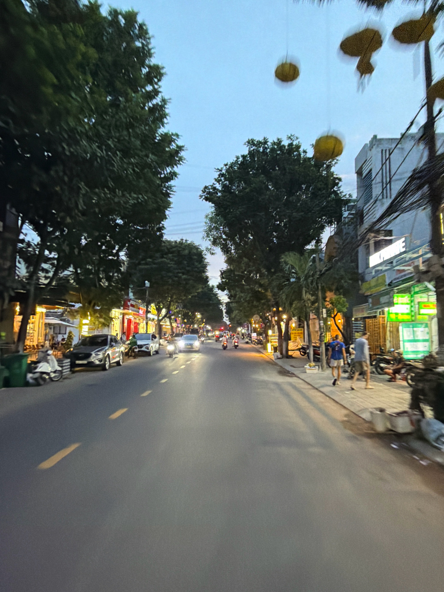 The image depicts a street scene in Da Nang, Vietnam, likely during the evening or twilight hours. The street is paved and has yellow lane markings. On the left side of the street, several cars are parked, including a silver SUV and a white sedan. A white scooter is also parked on the sidewalk. Large trees line the left side of the street, casting shadows. Buildings with illuminated storefronts are visible behind the trees. On the right side of the street, pedestrians are walking on the sidewalk. One person is wearing a blue t-shirt and shorts, and another is wearing a grey t-shirt and shorts. Several motorcycles are parked near the buildings. A few yellow lanterns are suspended in the air above the street. The sky is a pale blue, indicating the late hour.