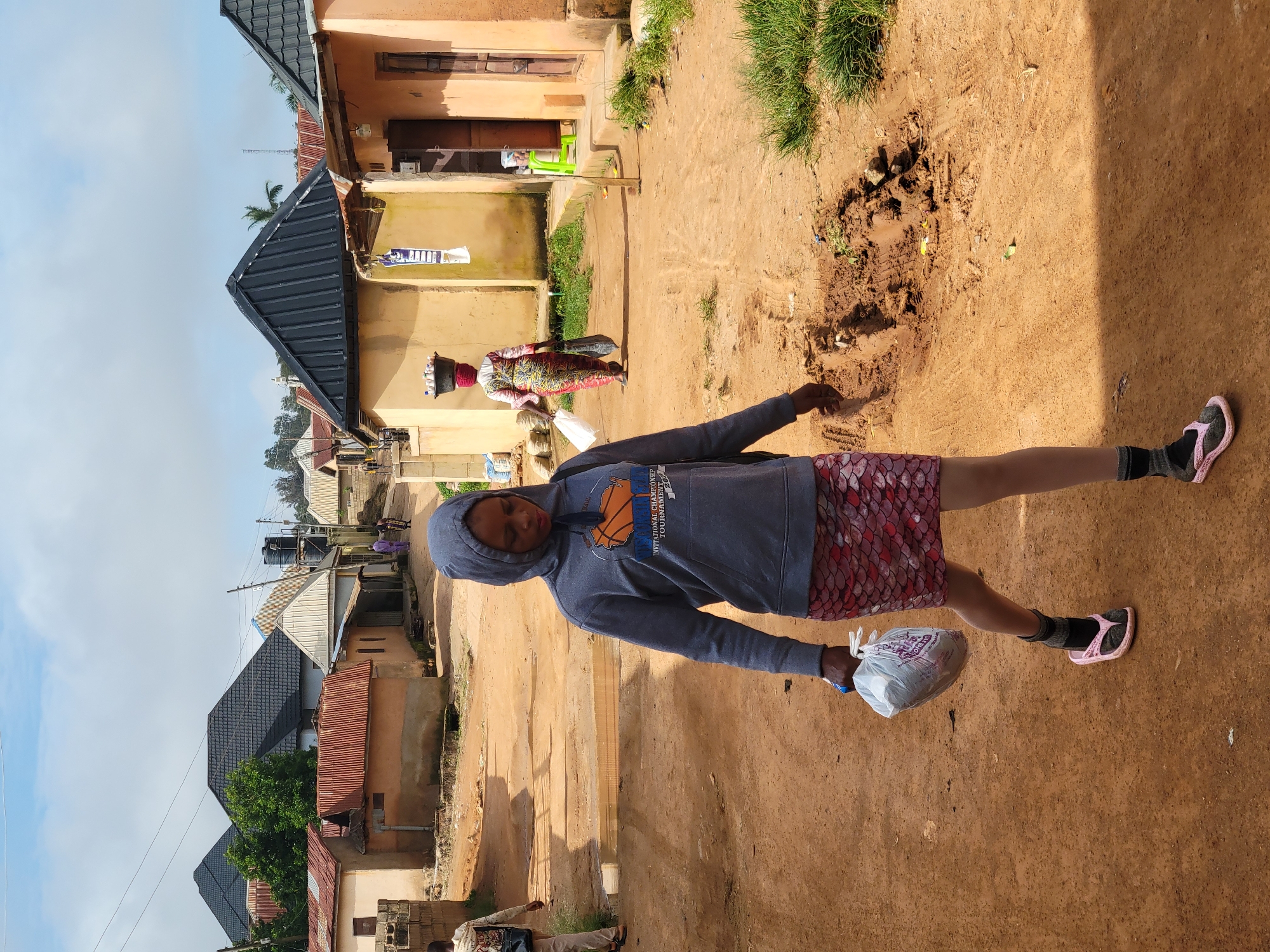 A woman wearing a gray hooded sweatshirt with an orange basketball graphic and a patterned skirt walks on a dirt road in Jos, Nigeria. She carries a white plastic bag in her left hand and a smaller, darker bag in her right. Behind her, another woman in a colorful patterned dress balances a basket of items on her head and carries a black bag. Several houses with corrugated metal roofs line the road. The sky is overcast.
