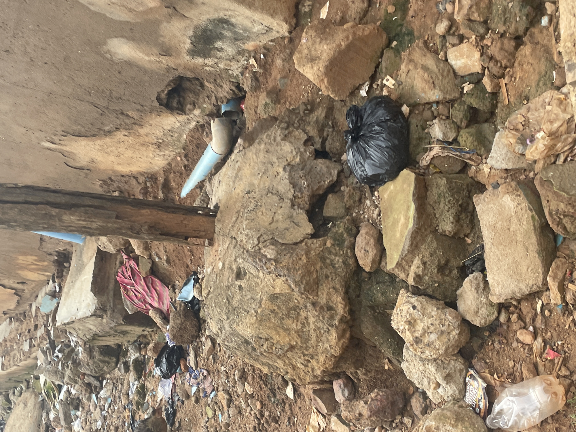 The image depicts a scene of urban decay in Lokoja, Nigeria. The ground is littered with rocks, rubble, and debris. Various discarded items are visible, including a red and pink patterned fabric, a black plastic bag, a blue pipe, and a clear plastic bottle with a brown liquid inside. A large, rough-textured rock dominates the center of the image, with smaller rocks and dirt surrounding it. In the background, a weathered brown wall with visible damage and holes suggests a dilapidated structure. A vertical wooden post stands prominently in the middle ground. The overall impression is one of neglect and unsanitary conditions.