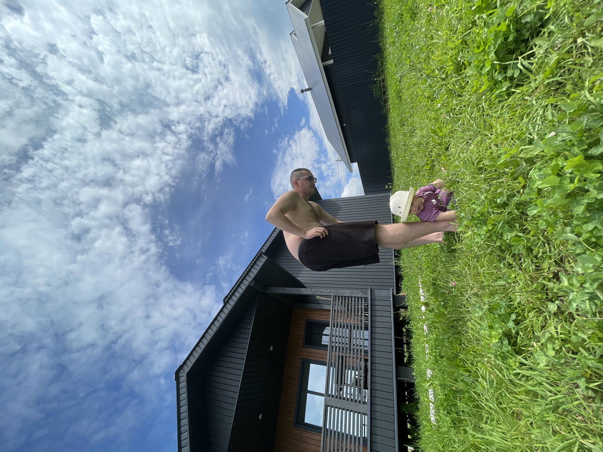 A man and a baby are on a grassy area in front of a dark-sided modern house with a wooden accent on the second story and a wooden deck with railings. The man is wearing dark shorts and is bent over, looking down at the baby. The baby is wearing a light-colored hat and a purple shirt, and is sitting on the grass. The sky is blue with white, wispy clouds. The location is in the village of Podshivalovo, Russia.