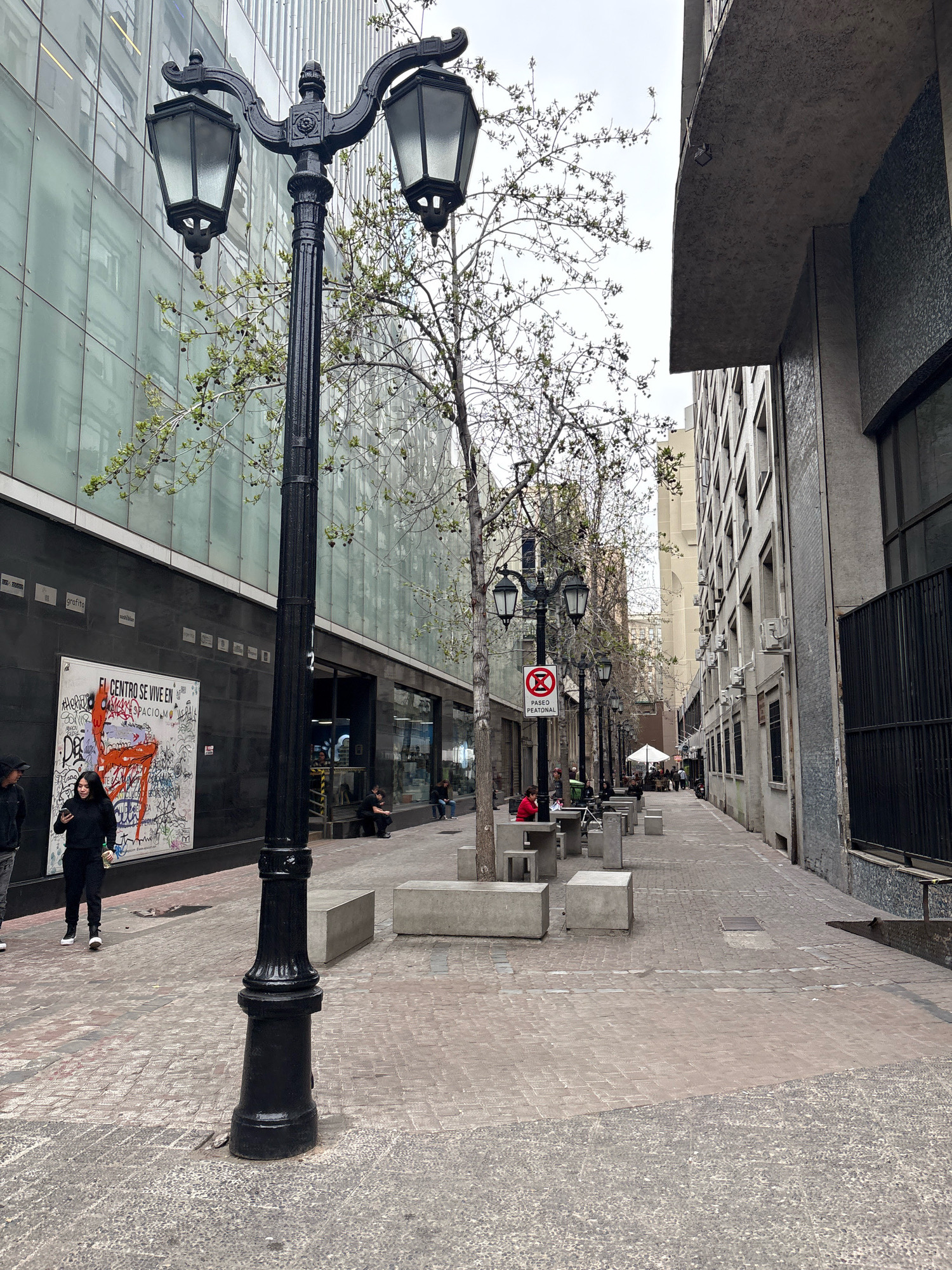This is a vertical, eye-level shot of a pedestrian alley in Santiago, Chile, on an overcast day. The alley is paved with a cobblestone-like pattern and lined with buildings. On the left, a modern building with a glass facade reflects the sky and surrounding architecture. Below the glass facade is a black wall covered in graffiti, including a large piece with orange spray paint. A black lamppost with two decorative lights stands prominently in the foreground on the left. A young woman wearing black is walking away from the camera in the foreground, looking at her phone. Further down the alley, there are several concrete benches and tables, with a few people sitting and walking. On the right, a concrete building rises up, featuring numerous window air conditioning units. At the far end of the alley, a white umbrella shades an outdoor seating area. A black metal fence runs along the right side of the alley near the foreground. The trees lining the alley are bare, indicating it is likely spring or autumn.