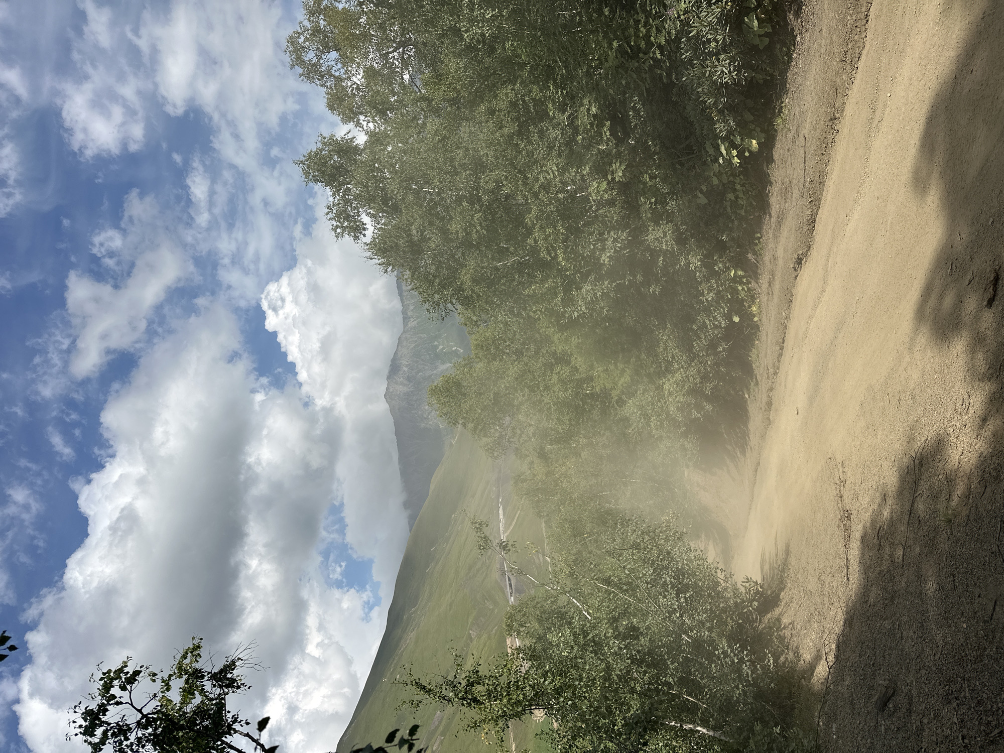 The image shows a dusty, unpaved road winding through a natural landscape. The road is a light brown color, suggesting it is made of dirt or gravel. On either side of the road, there are dense green trees and foliage. In the background, large, green mountains are visible, and the sky above is a mixture of blue with scattered white clouds. A noticeable cloud of dust hangs in the air above the road, indicating recent activity, possibly from a vehicle. The overall scene depicts a rural or mountainous environment, likely in a location with unpaved infrastructure. This image is not located in Lunnaya Polyana, Russia, as that city is a residential area and does not resemble this natural landscape.