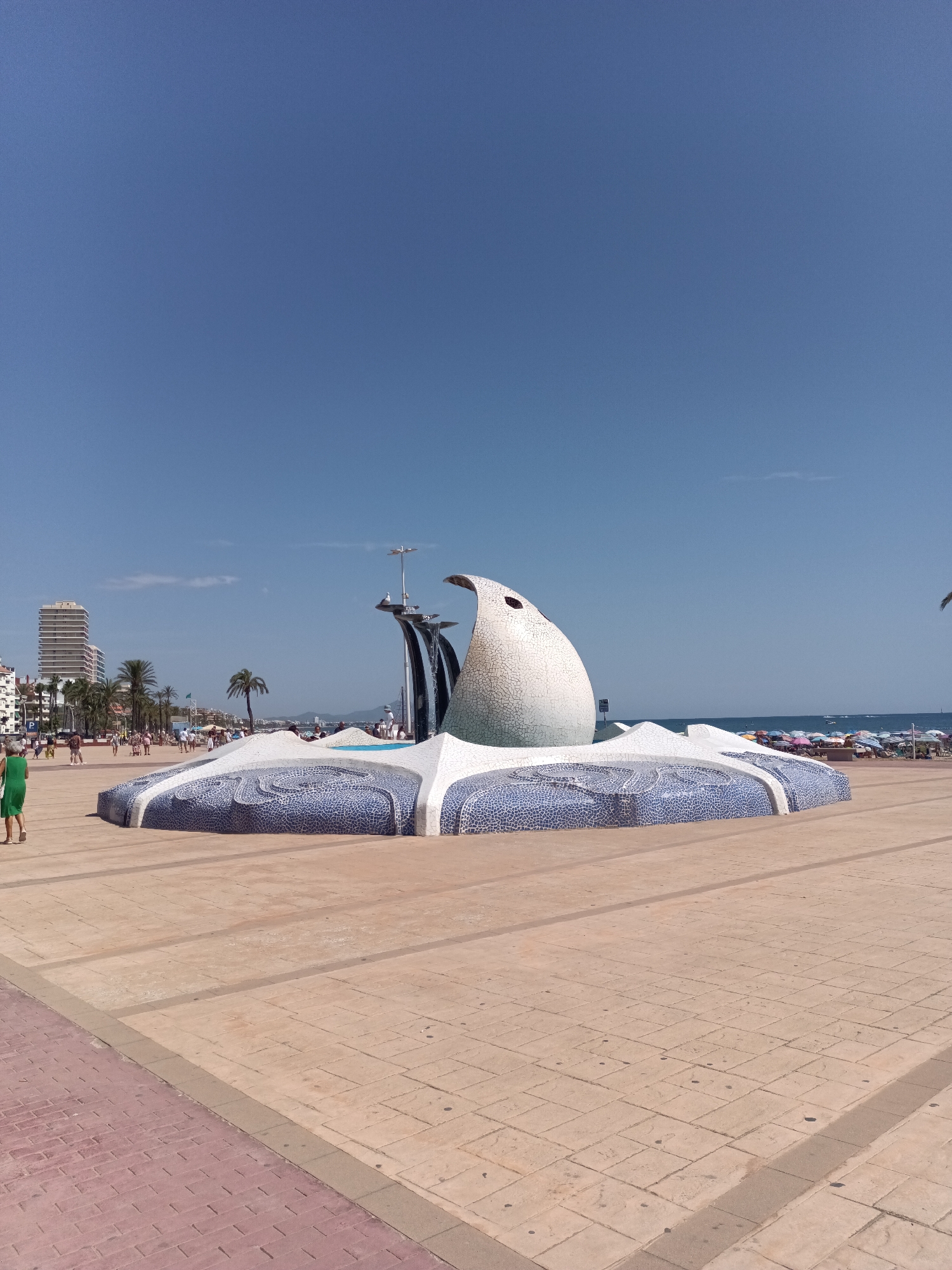 The image captures a waterfront promenade in Peníscola, Spain, under a clear blue sky. In the foreground, a large, wave-like fountain sculpture, adorned with blue and white mosaic tiles, dominates the scene. To the left of the fountain, a person in a green dress walks away from the camera. Behind the fountain, the Mediterranean Sea stretches to the horizon, dotted with numerous colorful beach umbrellas and a few distant boats. Along the coastline, palm trees and modern buildings are visible, characteristic of a coastal resort town. The promenade itself is paved with light-colored rectangular tiles.