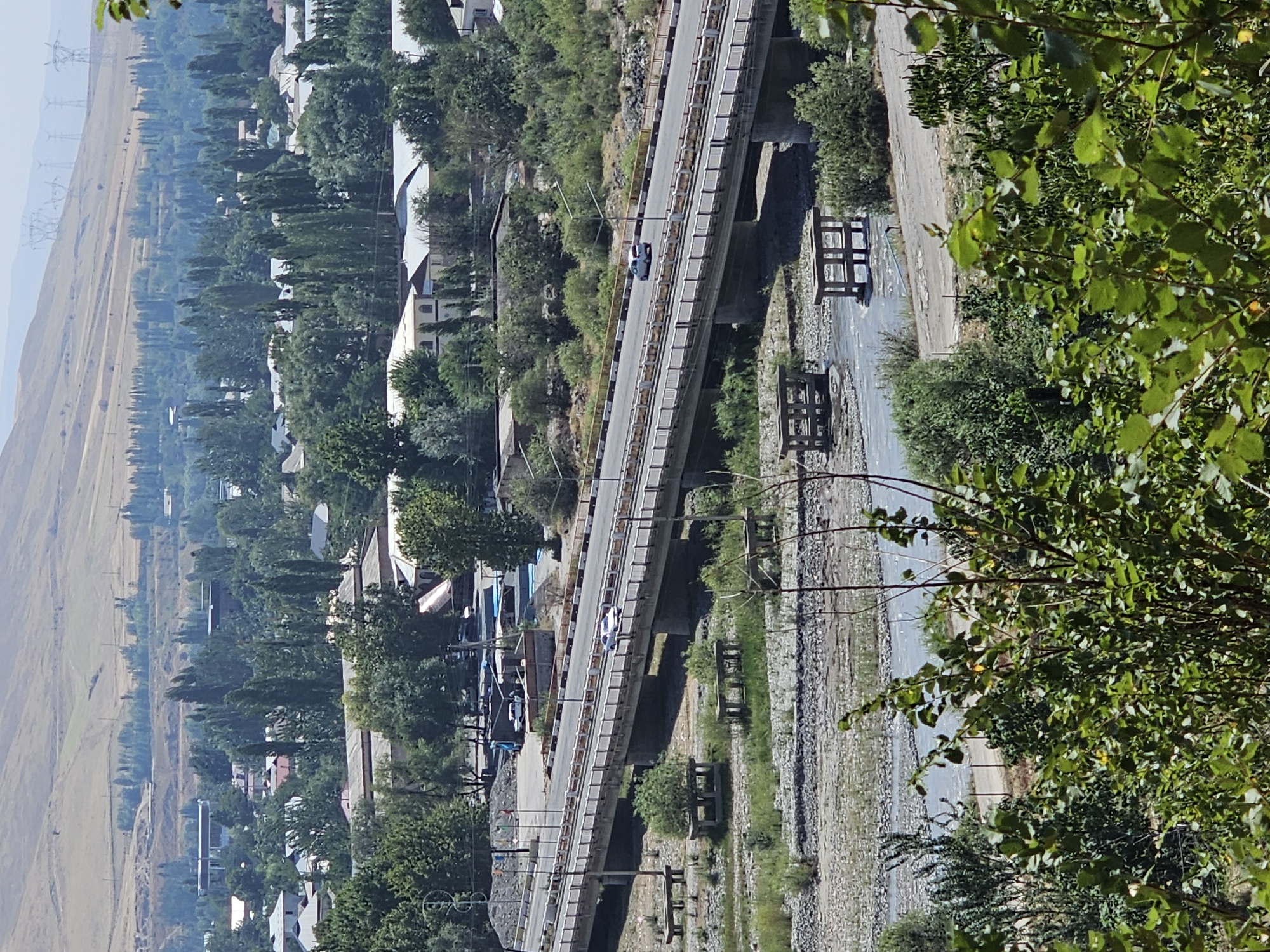 The image depicts a wide, elevated view of a city in Шералы, Kyrgyzstan. A long, multi-lane bridge spans a dry riverbed filled with rocks and a shallow stream. The bridge has a white railing and yellow accents along its edges. Several vehicles, including a white car, are visible on the bridge. Below the bridge, there are concrete structures and benches. The surrounding landscape features a dense collection of trees, houses with white roofs, and some industrial buildings. In the background, a dry, hilly terrain rises, dotted with power lines and transmission towers. The overall scene suggests a sunny day with clear visibility.