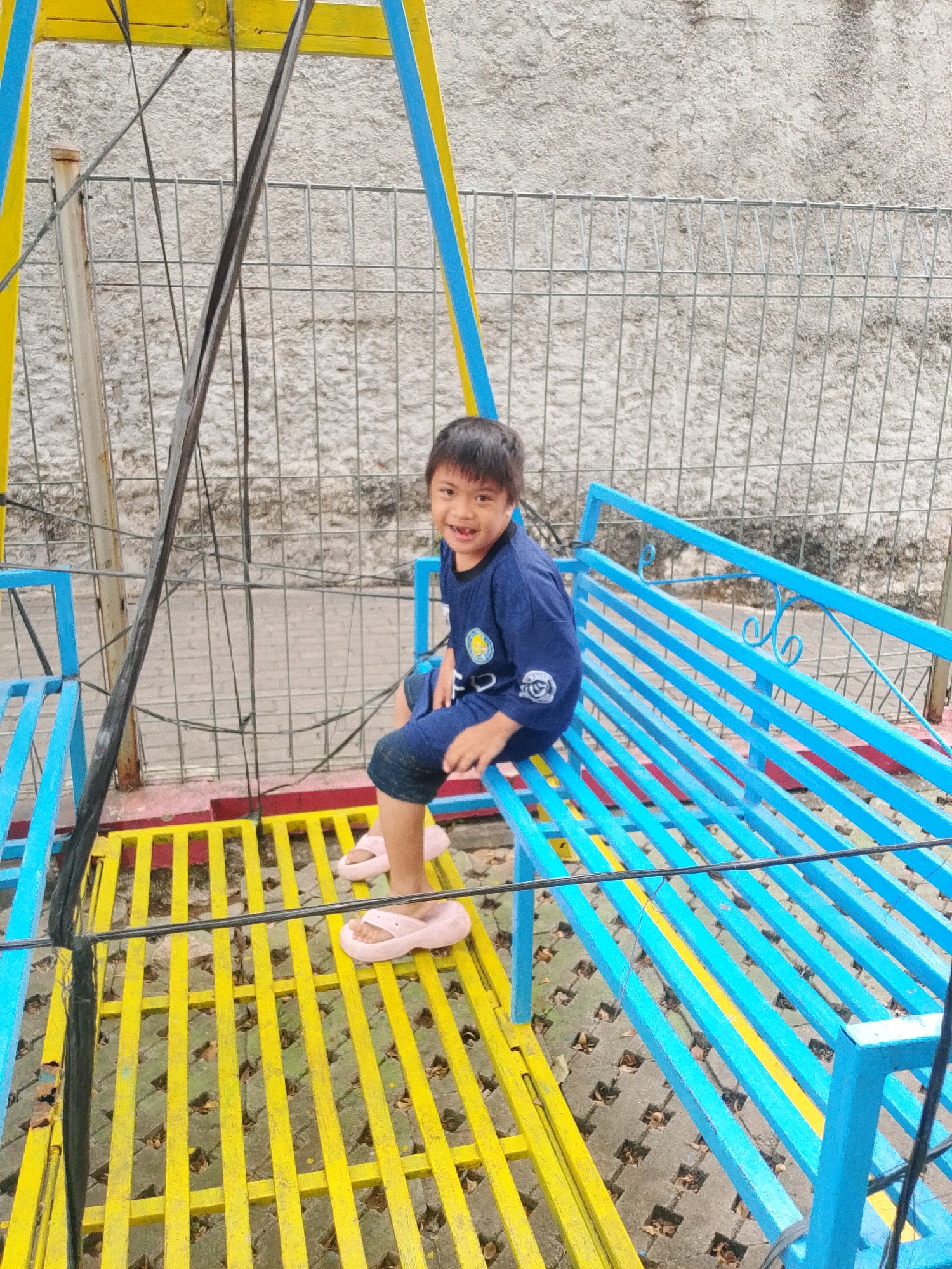 A young boy with dark hair is sitting on a blue bench of a swing set. He is wearing a navy blue t-shirt with a yellow emblem and dark blue shorts. The swing set is painted in bright yellow and blue colors, with a metal fence in the background. The ground beneath the swing set is paved with grey square tiles and scattered with fallen leaves. The boy is smiling at the camera. The scene appears to be an outdoor playground. The location is indicated as East Jakarta, Indonesia.