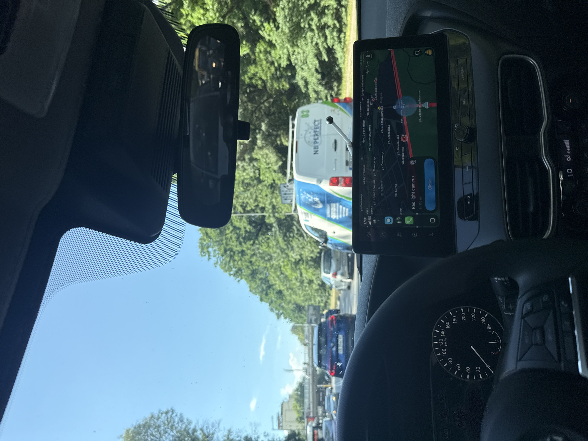 The image is taken from the interior of a vehicle, looking forward through the windshield and at the dashboard. The vehicle appears to be in traffic, with other cars visible on the road. The dashboard features a digital instrument cluster showing speed (approximately 20 km/h) and a GPS navigation screen displaying a map of Sofia, Bulgaria. The navigation screen indicates a "Red light camera" alert approximately 110 meters ahead. Outside the car, there are trees and a clear blue sky with some clouds. A white van with blue and green graphics and the text "NB PERFECT" is visible in front of the car. The image quality suggests it was taken with a smartphone camera.
