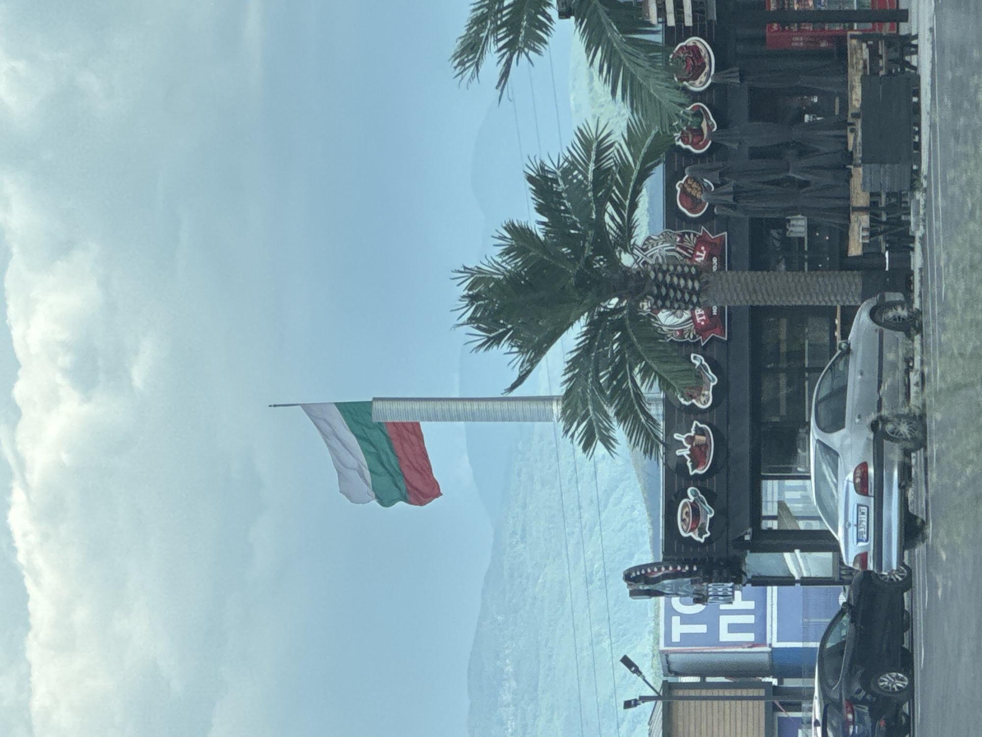 The image captures a street scene in Plovdiv, Bulgaria, marked by a blend of national symbols and commercial elements. A Bulgarian flag flies prominently on a pole, its white, green, and red stripes distinct against a backdrop of overcast sky and distant, hazy mountains. A palm tree stands nearby, suggesting a more temperate or stylized representation. Adjacent to the flag and palm tree is a building featuring prominently displayed signage and decorative elements, potentially indicating a restaurant or establishment catering to tourists. Cars are parked alongside the curb, typical of urban areas. The overall scene suggests a location of interest, possibly a tourist destination or commercial district within Plovdiv.