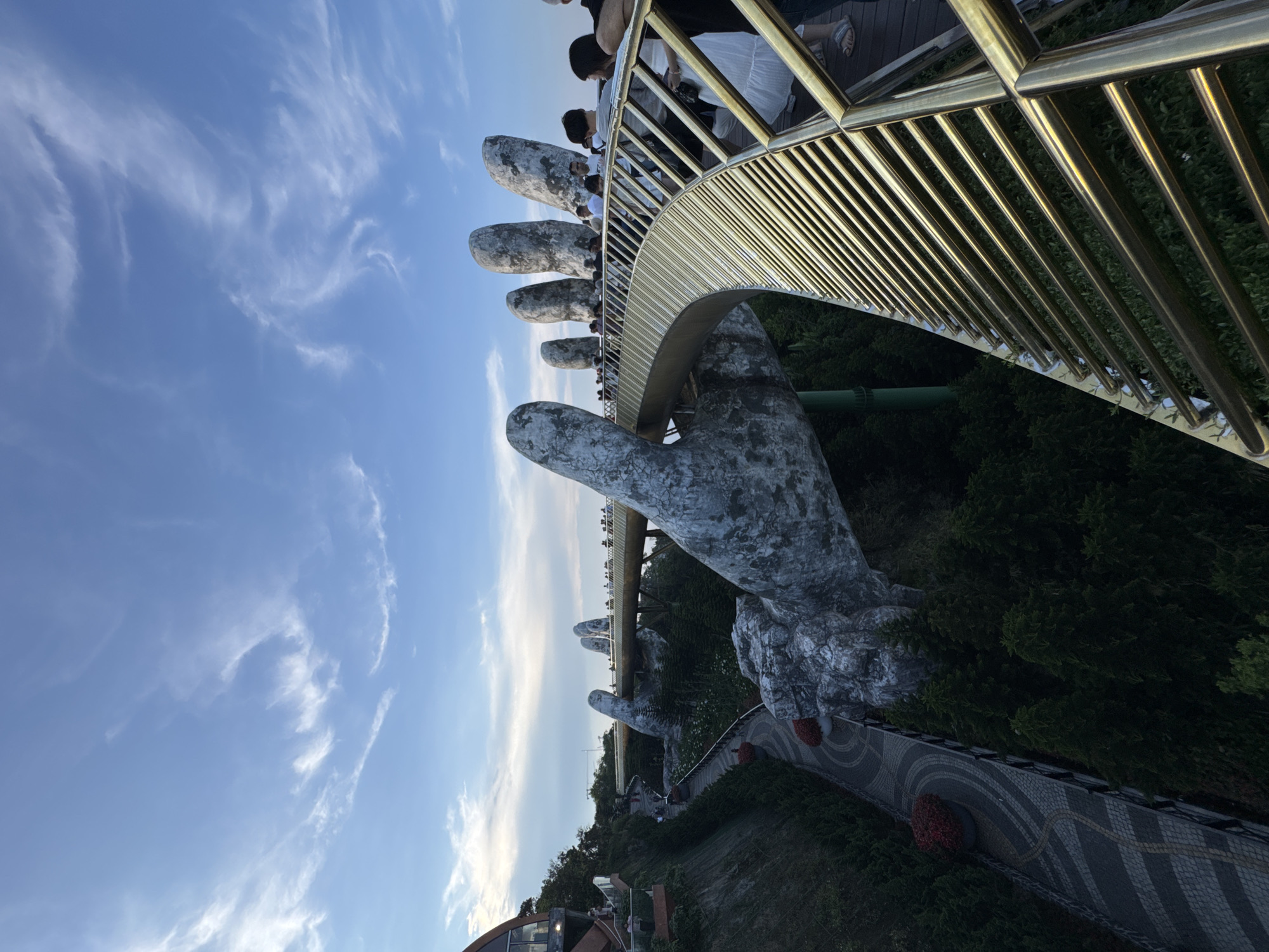 The image displays the Golden Bridge (Cầu Vàng) at Bà Nà Hills, located near Đà Nẵng, Vietnam. The scene is captured from an elevated perspective, showcasing the distinctive pedestrian bridge with a golden-colored metal handrail and a dark deck. The bridge is structurally supported by two colossal, weathered grey hands, sculpted to appear as if emerging from the mountainous landscape.
Numerous individuals, presumed to be tourists, are present on the bridge, walking and observing the surroundings. The bridge curves gently towards the horizon, where more people are visible. Below and surrounding the bridge structures, dense green foliage, consisting of trees and shrubs, covers the steep terrain. A winding, paved pathway with geometric patterns and circular planters containing red flowers is visible through the greenery at a lower elevation. On the far left, a modern building with a curved, dark-paneled and glass facade is partially visible, built into the hillside. The sky above is light blue with scattered, wispy white clouds, indicating fair weather. The lighting suggests late afternoon or early evening.