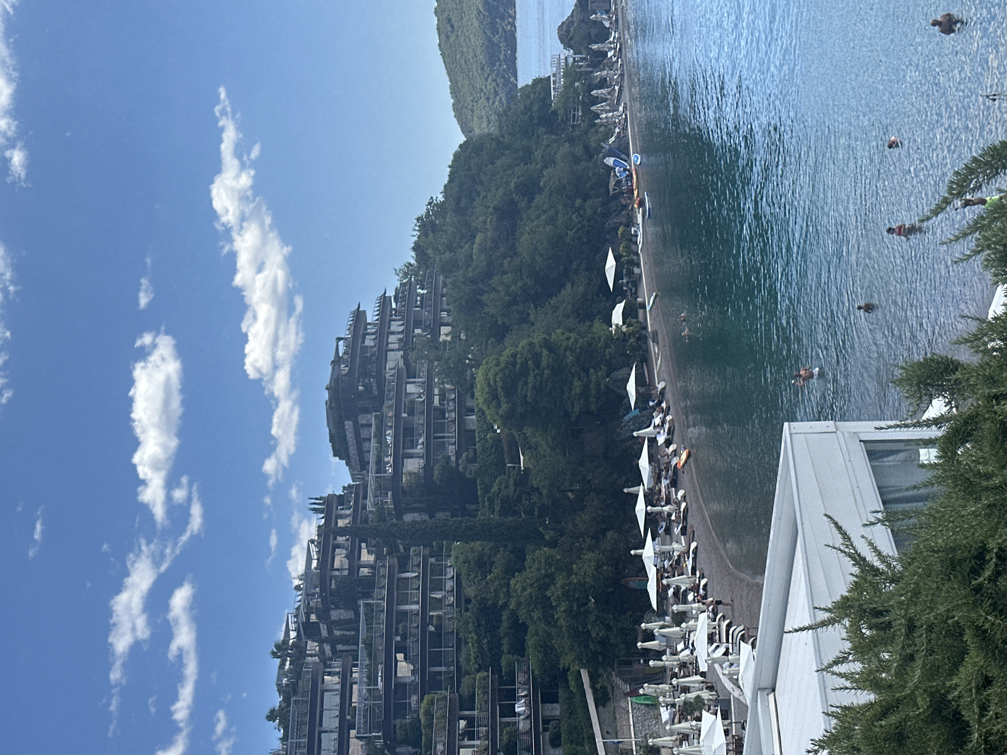 This image shows a resort complex built into a hillside overlooking a beach and the Adriatic Sea, likely in or near Budva, Montenegro. The resort features multiple levels of balconies and terraces, covered in greenery, cascading down the slope. Below the resort, a dark pebble beach is lined with numerous white umbrellas and lounge chairs. Several people are visible on the beach and in the water. The sea is a vibrant blue, with a clear sky dotted with white clouds above. In the foreground, part of a white building and lush green foliage are visible.
