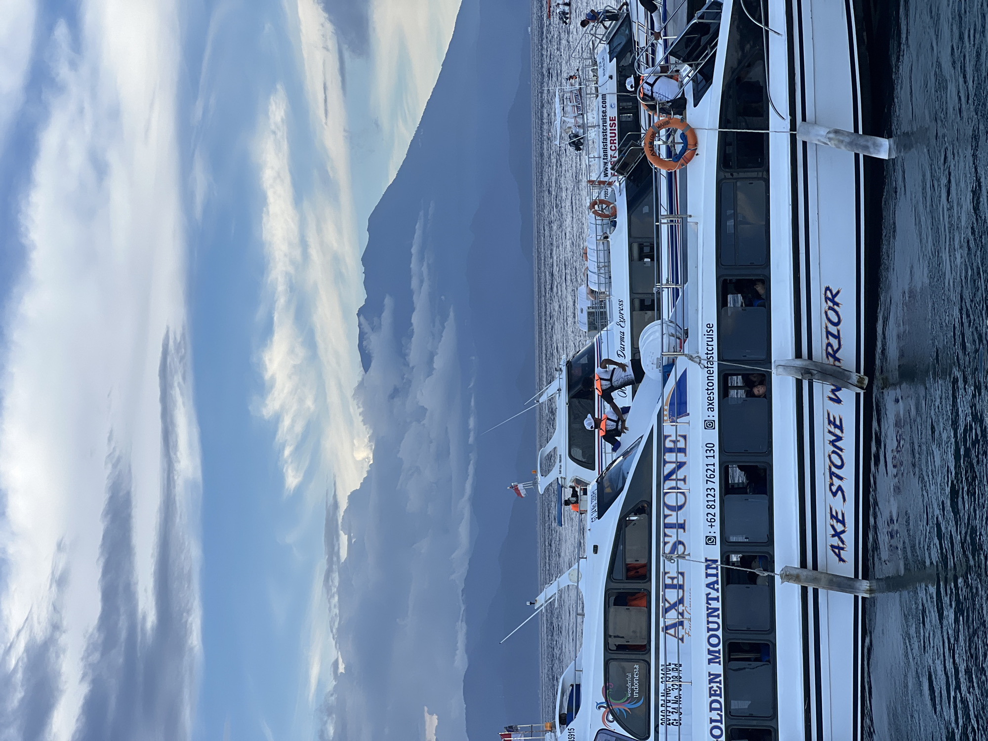The image captures a scene in Kampung Toyapakeh, Indonesia, featuring several white passenger boats docked near a mountainous shoreline. The boats, marked with the text "AXE STONE" and "GOLDEN MOUNTAIN", display contact information including a phone number (+62 8123 7621 130) and Instagram handle (@axestonefastcruise). Additional text mentions "Darma Express" and "tanisfastcruise.com" suggesting related services or branding. People can be seen inside the boats, some wearing orange life vests. The background includes a mountain range partially obscured by clouds. The sky is characterized by a mix of blue and grey clouds. The boats are aligned along a dark, calm body of water. Wooden posts are visible along the boats, used for mooring.
