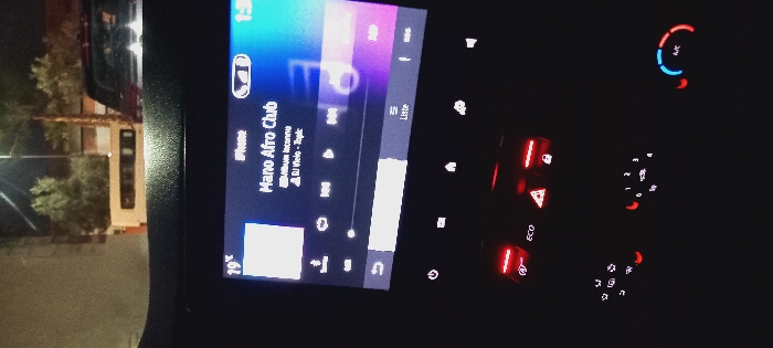 An in-car entertainment system displays "Mano Afro Club" by "Album Inconnu" and DJ Velo - Topic. The system's display indicates a temperature of 19°C. A car air conditioning system is visible, and other dashboard buttons with red accents are illuminated. A glimpse of the outside world, showing trees and buildings, is visible through the car's window.