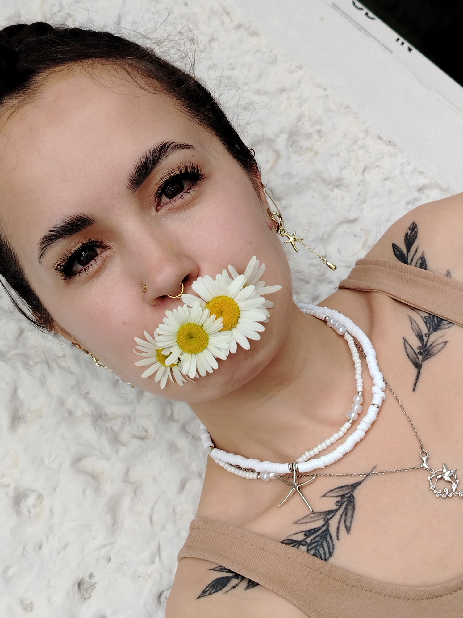 A close-up, high-angle medium shot shows a young woman with dark hair pulled back, lying on a textured white surface. She is positioned with her head angled slightly to her left, and her eyes are looking towards the camera. Three white daisies with yellow centers are held in her mouth. She has a gold septum piercing and a small gold hoop earring in her left ear. She is wearing a light brown tank top. There are several black tattoos on her upper chest and shoulder, depicting leafy branches. She is wearing two necklaces: one with white disc beads and pearls, and another longer silver chain with a starfish charm and a hummingbird charm. The background is a rough, textured white surface, possibly a wall or ground. The overall impression is one of a stylized portrait. The image does not contain information about its location in Noviy, Russia.