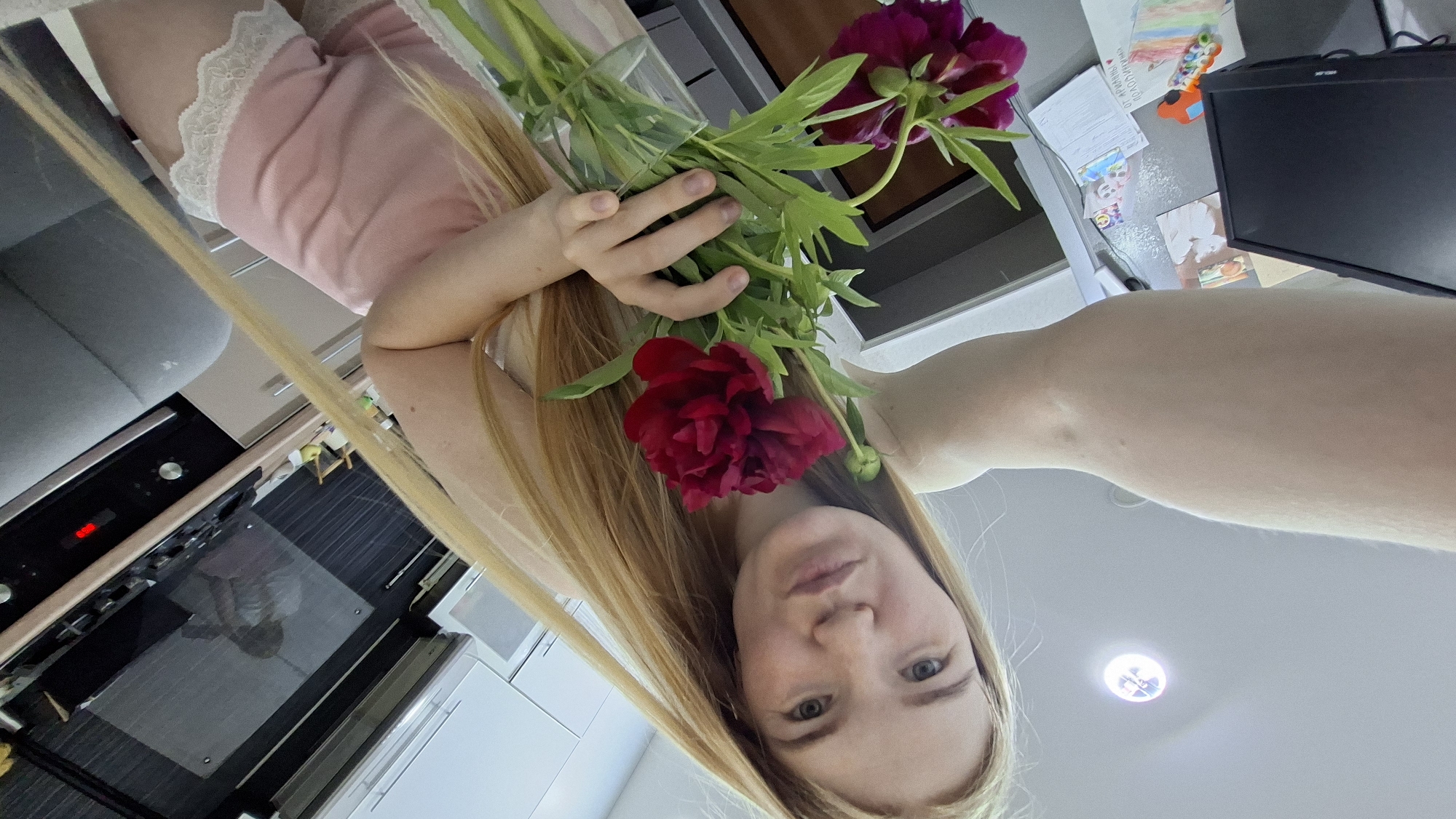 The image captures a unique overhead selfie perspective of a young woman in an indoor domestic setting, identified as Chaikovsky, Russia. The woman, with long, straight blonde hair and a fair complexion, looks directly into the camera with a neutral, serene expression. She holds a vibrant bouquet of dark red peonies with green stems and leaves, positioned near her face.
The scene appears to be a kitchen or a combined living and kitchen area. To the left, a gray sofa or chair is partially visible. Below it, a black oven with a glass door reflects parts of the room and the woman's lower body, revealing she is wearing light pink shorts with white lace trim. A red light indicator is visible on the oven. To the right, a kitchen counter or table holds a black flat-screen computer monitor, partially displaying the brand "AOC." Scattered on the counter are several papers, including colorful children's drawings and sheets with handwritten Cyrillic text, some partially legible such as "ПРО...О...", alongside other illegible markings. White kitchen cabinets are also visible in the background and reflections. A circular recessed spotlight on the ceiling provides overhead illumination. The image depicts a casual, personal moment at home, likely taken during the daytime or evening with artificial lighting.