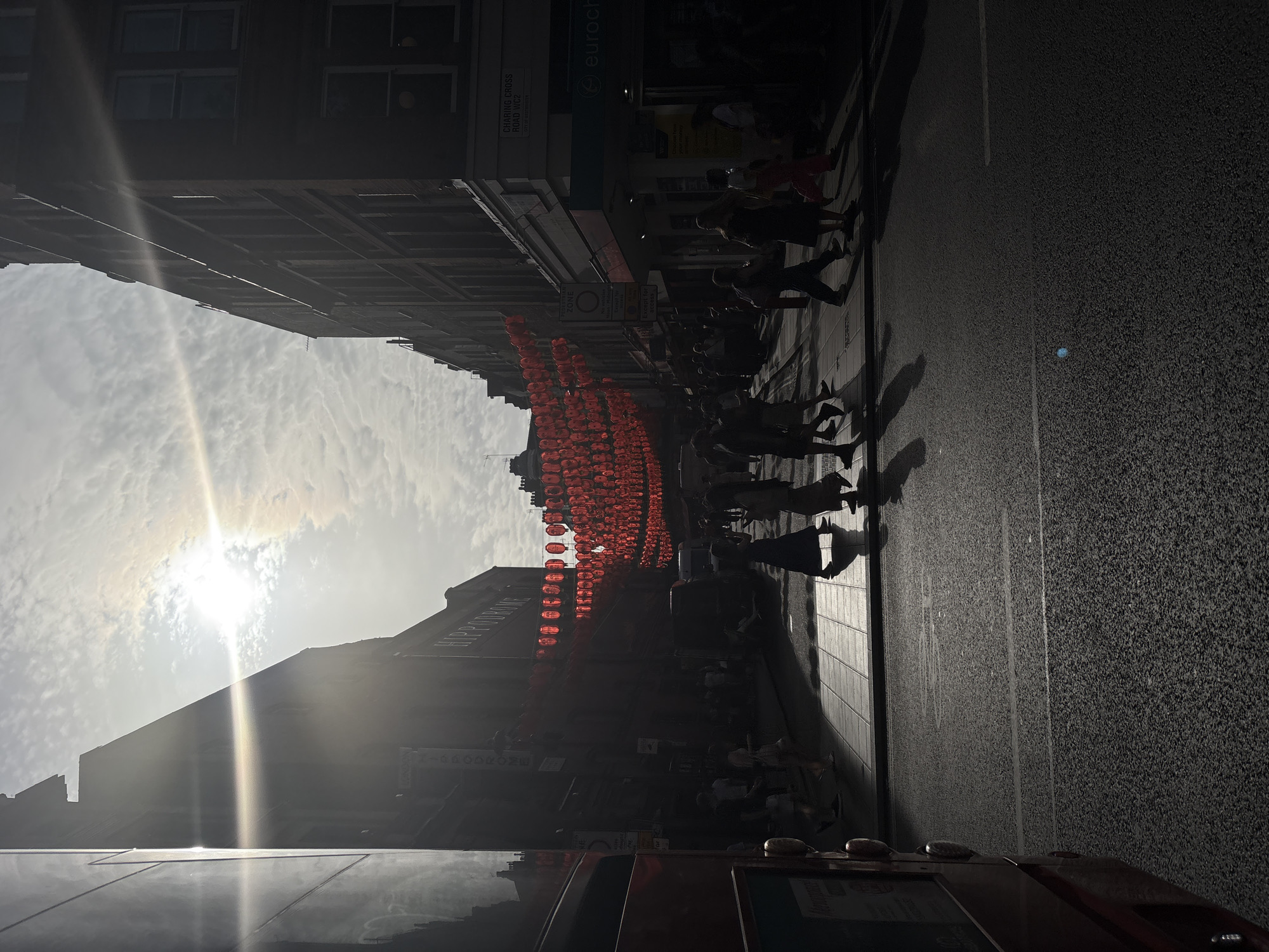 This image shows a street scene in Chinatown, London, with red lanterns strung across the street. People are walking along the sidewalk. The sky is bright, with the sun partially obscured by clouds. Buildings line the street, casting long shadows. The image seems to be taken from the top deck of a bus, looking down at the street. The road is empty with gray asphalt.