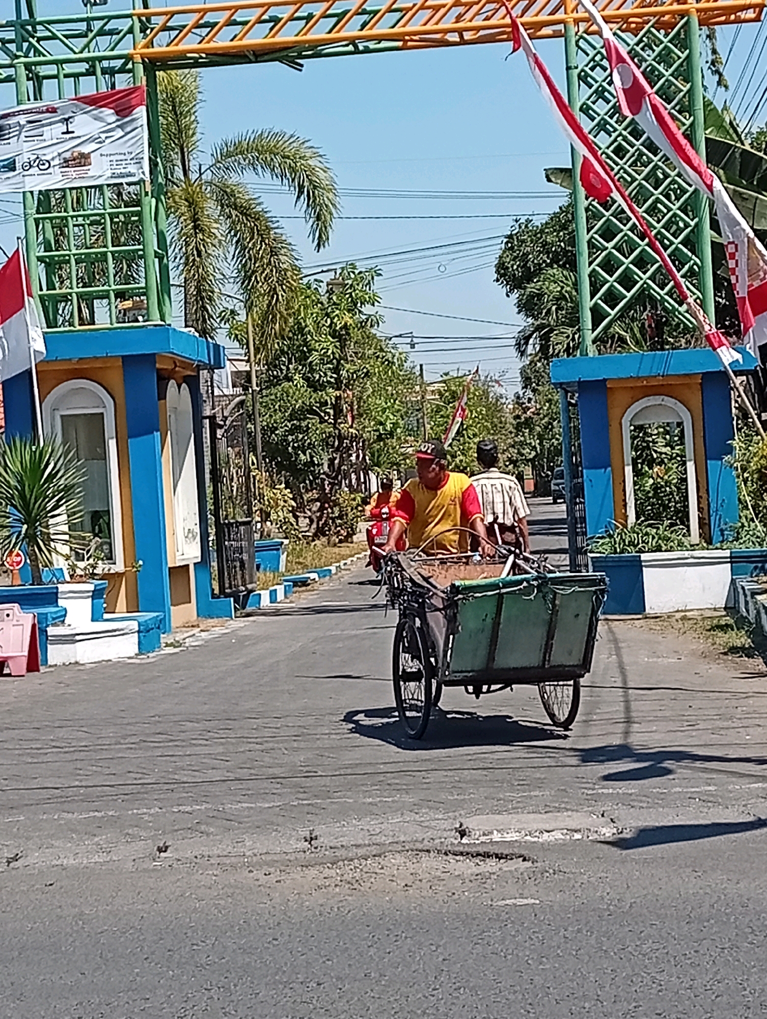 An outdoor, medium shot captures a bright, sunny day in Sidoarjo, Indonesia, focusing on a man riding a bicycle cart on a paved road. The man, wearing a yellow and red shirt and a red baseball cap, is in the foreground, pedaling the cart, which is filled with green crates. Behind him, another man in a striped shirt walks on the right side of the road. The road appears to have some minor damage, with small patches and cracks visible.
The scene is framed by an archway decorated with green metal structures and yellow bars, adorned with red and white Indonesian flags, suggesting a festive or national occasion. On the left side of the archway, a large banner with text and images is displayed. Palm trees and other lush green foliage are visible behind the archway and along the sides of the road, interspersed with residential-style buildings. Two blue and yellow structures with arched doorways stand on either side of the road beneath the archway. Power lines are present overhead. The overall atmosphere is that of a typical Indonesian street scene under clear blue skies.