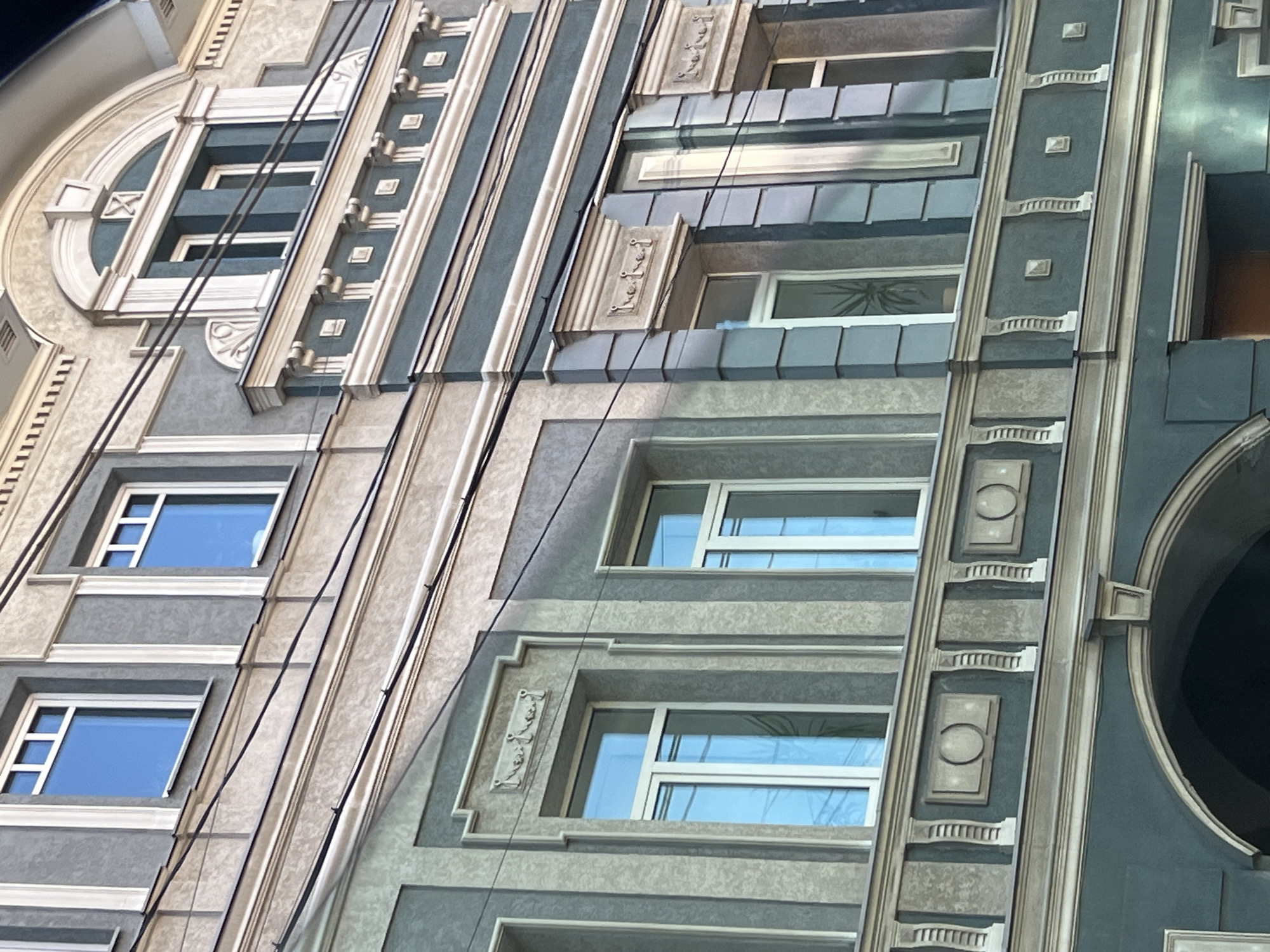 The image shows a close-up view of the facade of an old building in Nizhny Novgorod, Russia. The building exhibits neoclassical architectural elements with a grey and teal color scheme. There are several windows with white frames, some of which reflect the blue sky. Decorative plasterwork, including moldings and relief ornaments, adorns the facade. Wires are visible running across the building's exterior. The lower part of the image reveals an arched entrance. The overall impression is of a historic urban structure.
