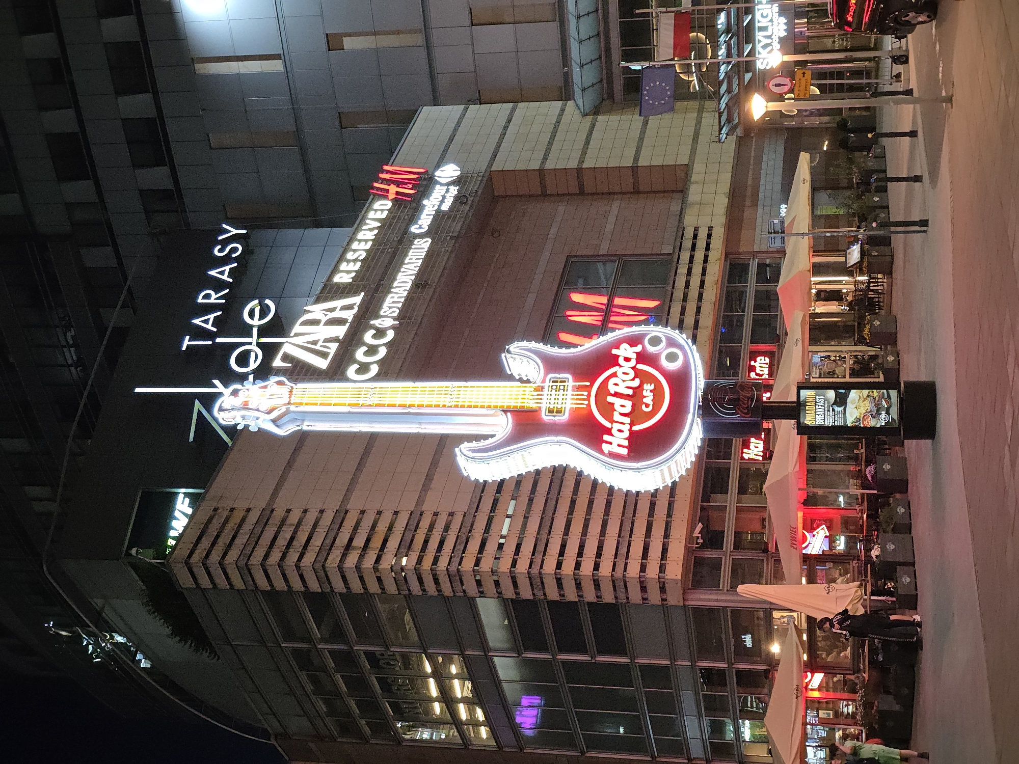 The image depicts a night scene in Warsaw, Poland, featuring a Hard Rock Cafe. A large, illuminated guitar sign of the Hard Rock Cafe stands prominently in front of a modern building. The building has multiple floors with glass windows and facades, showcasing various brands like Zara, Reserved, CCC, Stradivarius, Carrefour market, and Tarasy.
Several people are visible near the entrance of the Hard Rock Cafe, some walking, suggesting it is a lively area. Adjacent to the building is an outdoor dining area with tables and chairs, covered by white umbrellas, likely belonging to the cafe. Flags, including the European Union and Polish flags, are displayed on poles near the top of the building. Street lamps illuminate the scene, casting light on the building and the surrounding pedestrian area.