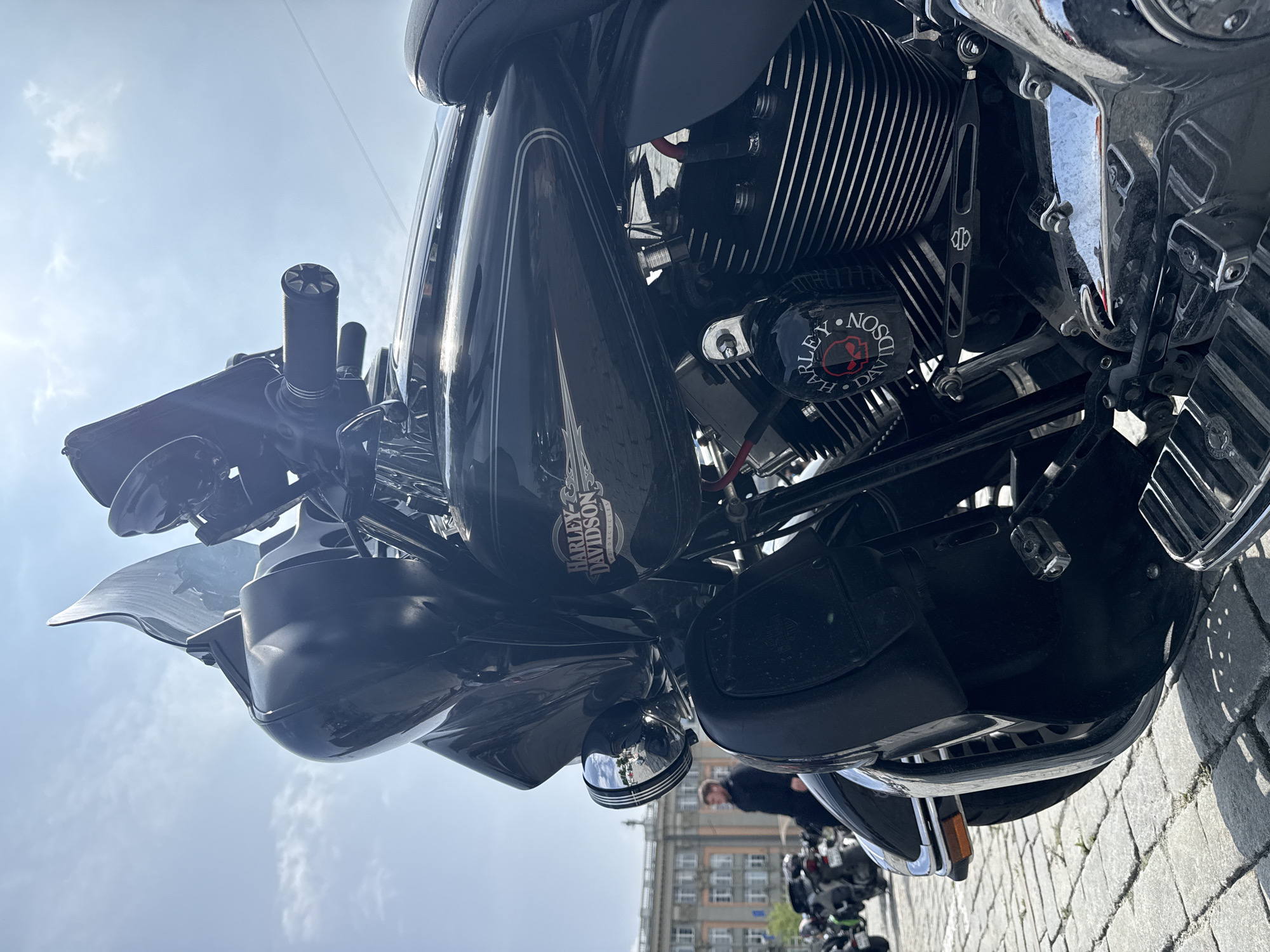 A low-angle, close-up photograph captures the front and right side of a black Harley-Davidson motorcycle parked on a cobblestone surface under a bright, partly cloudy sky in Yekaterinburg, Russia. The motorcycle features a large black fairing with a tinted windshield, handlebars with textured black grips, and a black fuel tank with "HARLEY-DAVIDSON" branding and silver pinstriping. The visible V-twin engine has black finned cylinders and a distinctive black air cleaner cover with a red skull and "HARLEY DAVIDSON" text. Chrome accents are visible on the engine casing, exhaust pipe, and front fender trim. Black footboards are present on the right side. In the mid-ground, to the left of the motorcycle, a male figure dressed in a dark jacket walks past a large, light-colored historical building with multiple windows. Several other motorcycles are visible parked further in the background to the left. Sunlight illuminates the scene from the upper left, casting defined shadows on the cobblestones and creating strong reflections on the motorcycle's chrome surfaces.