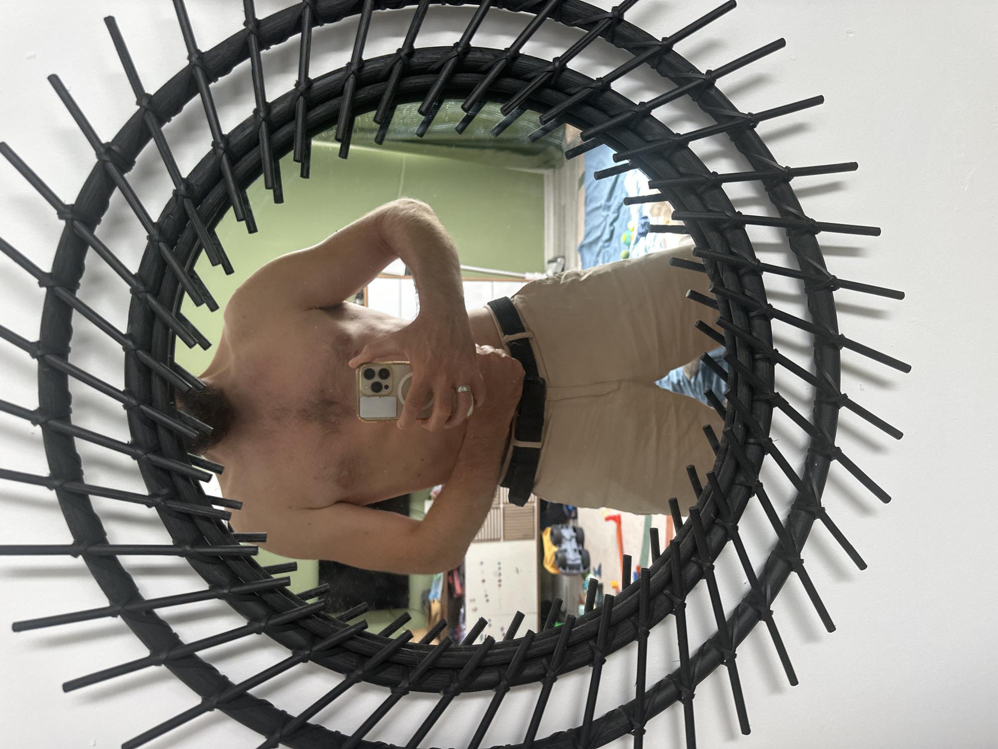 A person is taking a selfie in a round, black, rattan-framed mirror. The person is shirtless and wearing light-colored shorts and a black belt. They are holding a smartphone in their right hand. The background of the mirror reflection shows a room with furniture, toys on the floor, and a green wall. The scene is indoors, likely in a residential setting. There is no indication of the location being Fethiye, Turkey.