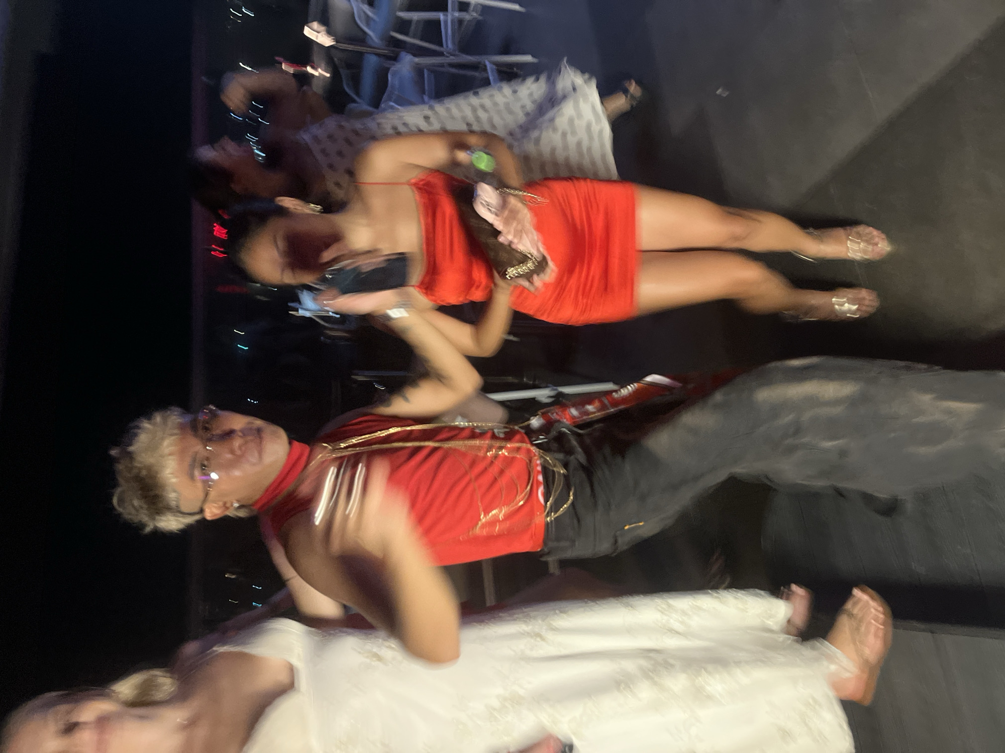 The media file captures a nighttime scene of several people gathered, likely at an outdoor event or party. The overall tone appears festive, with bright lighting and vibrant attire.
In the foreground, a person with blonde, styled hair and glasses is visible from the shoulders up. They are wearing a red garment with gold chain detailing. Below them, another individual is partially visible, wearing a white, possibly lacy, garment.
Further back, a woman in a red, form-fitting dress is the most prominent figure. She is holding a light-colored clutch bag and is wearing transparent, strappy sandals. Her pose and the way she is holding the bag suggest she is socializing or posing for a photo. Other individuals are present in the background, but are less distinct due to motion blur and lighting.
The setting appears to be a flat, dark surface, possibly a rooftop or outdoor patio, with some elements of furniture or staging in the background. The lighting is artificial and seems to be a mix of ambient light and perhaps stage lighting, creating a dynamic and lively atmosphere.
The presence of multiple people, their attire, and the overall ambiance suggest a celebratory event, possibly a party or gathering. The context of Đà Nẵng, Vietnam, does not provide specific visual cues within the image itself, but it anchors the location to a coastal city known for its nightlife and events. The time of day is clearly night due to the darkness of the background and the use of artificial lighting. No visible text is present in the image.