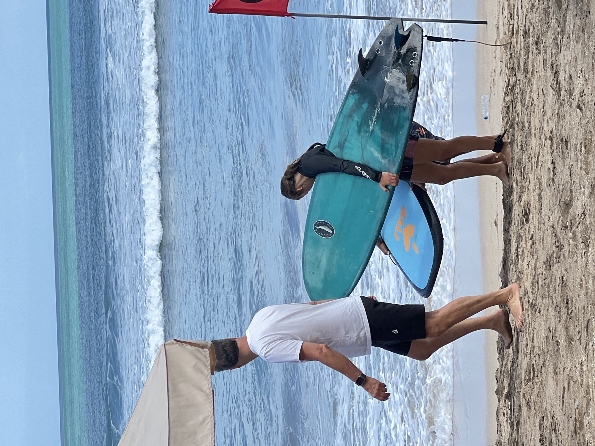 A man and two young individuals are walking on a sandy beach in Kuta, Indonesia, carrying surfboards. The man, wearing a white t-shirt and black shorts, walks towards the ocean. He has a watch on his left wrist. To his right, a young person in a black rashguard and shorts holds a turquoise surfboard. Another young person, partially visible, is carrying a smaller blue surfboard underneath the turquoise one. The ocean is visible in the background with small waves breaking. A red flag on a pole stands to the right of the scene. A small canopy is present on the left side of the frame.