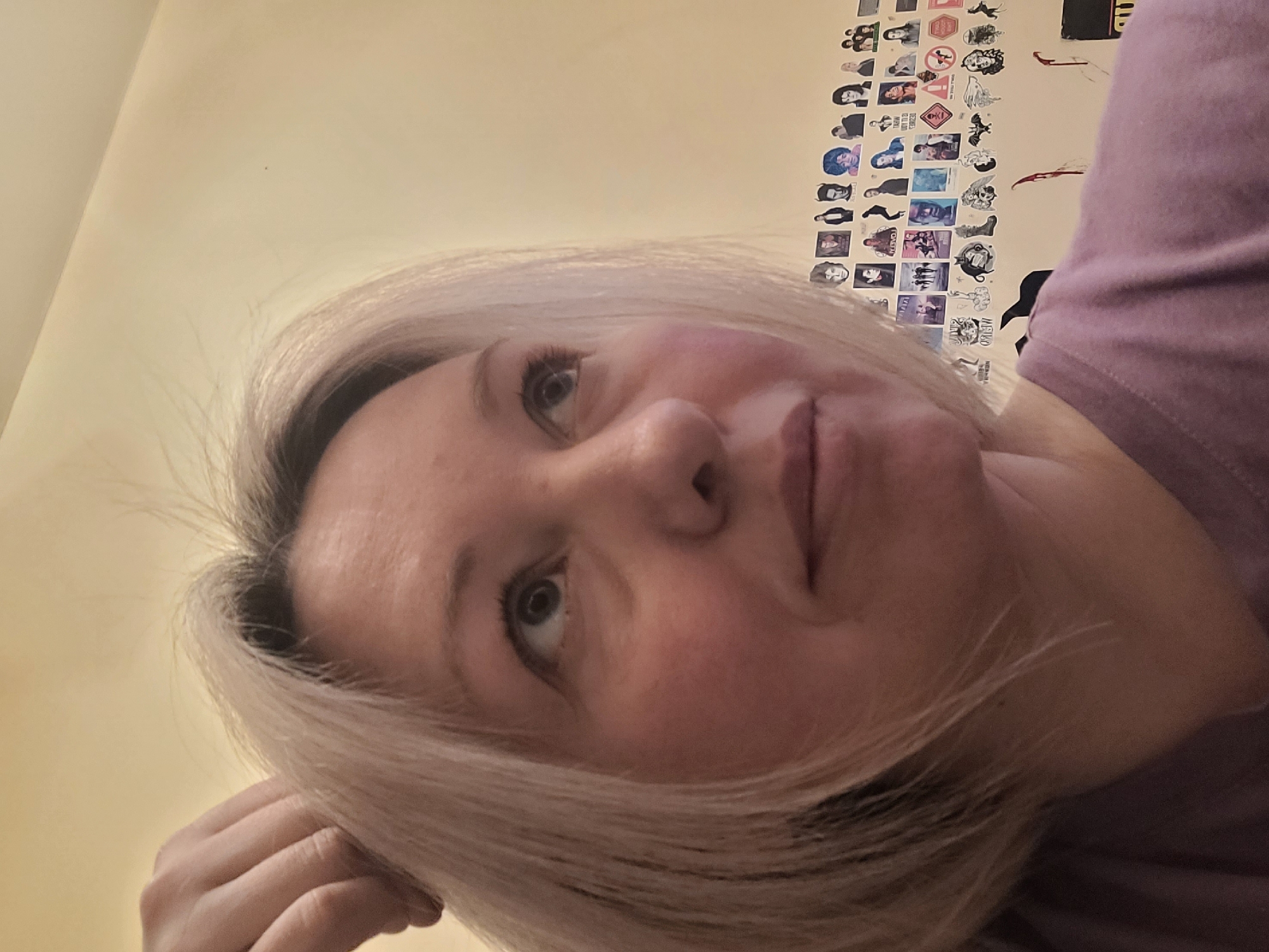 The image depicts a woman with blond hair, fair skin, and light-colored eyes, looking upwards towards the left. Her right hand is near her face, suggesting she might be resting her head on it. She's wearing a mauve-colored top. Behind her, the wall is a light yellowish color and covered with stickers and what appears to be photographs. There's no information in the provided image that would connect it to Izhevsk, Russia.