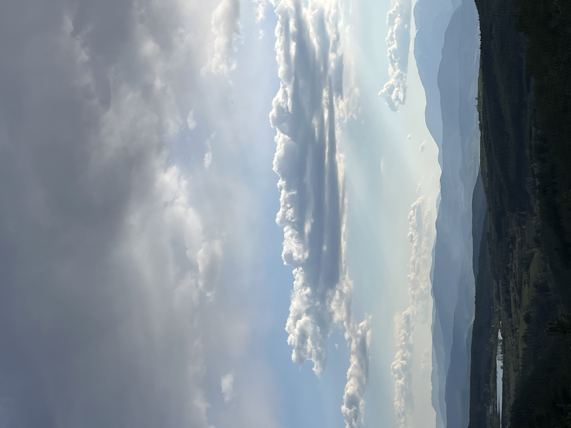 The image presents a landscape with mountains and a sky filled with clouds. On the right side of the image, layered mountain ranges are visible, gradually fading in color due to atmospheric perspective. In the foreground, the terrain is darker, showing hints of greenery and possibly forests. A body of water is also partially visible. The sky dominates the left side of the image, with a mix of dark, ominous clouds and brighter, sunlit clouds. Light appears to stream through some of the cloud formations. I do not have any information about Kribul, Bulgaria.