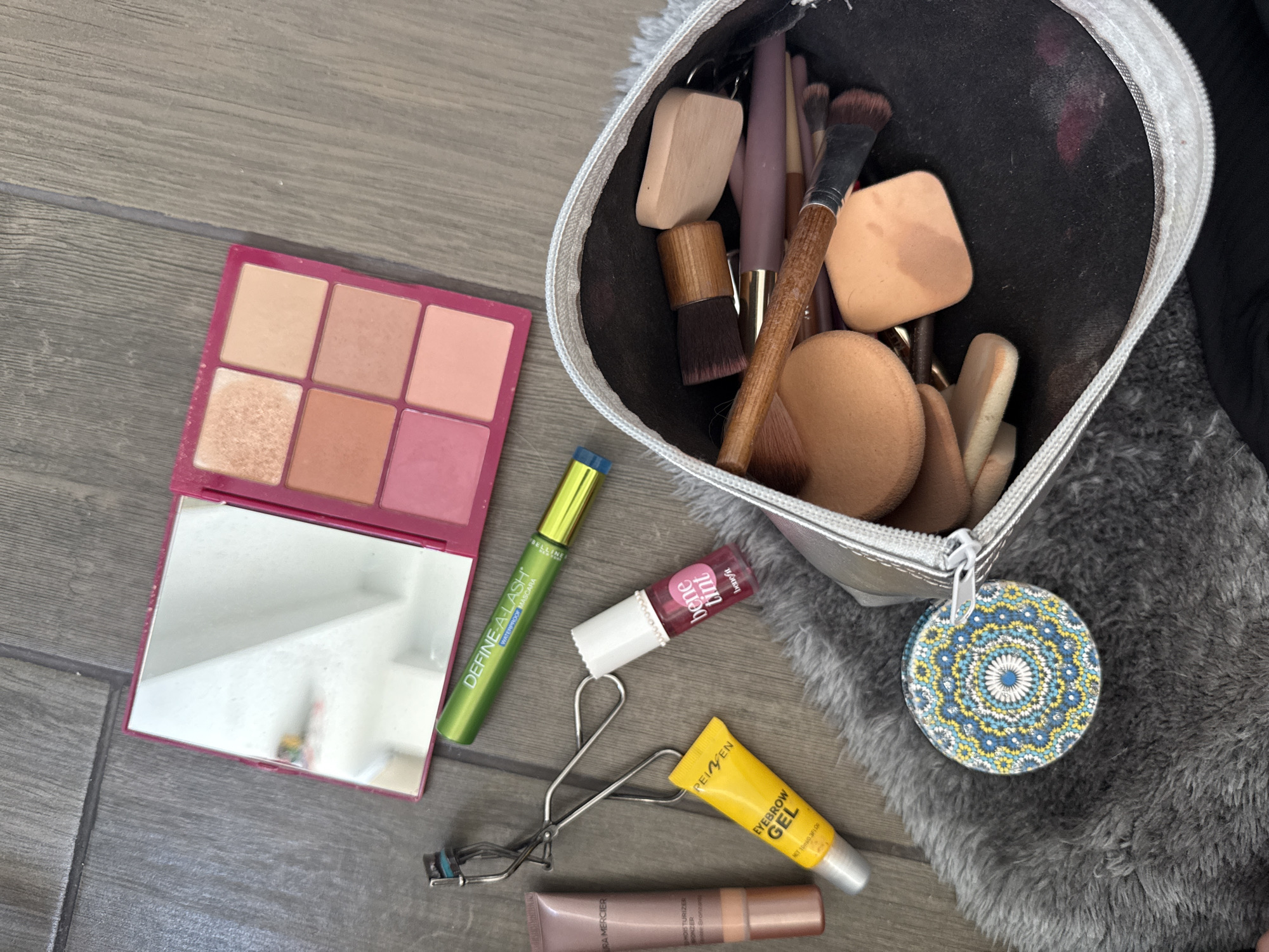 An overhead shot displays a collection of makeup products and accessories laid out on a surface primarily composed of wooden floorboards, with a grey furry rug covering part of the right side. The scene appears to be an indoor setting.
On the left, a vibrant pink rectangular makeup palette is open, revealing six square pans of blush or eyeshadow in shades of peach, coral, and pink, with one shimmery champagne color. The lower half of the palette contains a mirror reflecting a blurry indoor background.
To the right of the palette, various individual makeup items are scattered. A green tube of mascara, labeled "DEFINE-A-LASH WATERPROOF MASCARA," is prominent. Below it, a metallic eyelash curler rests alongside a small white tube of Benefit "bene tint," a lip and cheek stain. Further down, a yellow tube of "PEI'EN EYEBROW GEL" is visible. The final product in this arrangement is a rose gold tube, identified as "URA MERCIER MOISTURIZER BONZER" (likely Bronzer).
On the right side of the image, a silver-trimmed, dark-colored makeup bag stands open on the grey furry rug. It is filled with an assortment of makeup tools, including several brushes with wooden and synthetic handles, and numerous beige blending sponges of various shapes and sizes. Some red cosmetic smudges are visible inside the bag. A circular, ornate keychain or charm, featuring a blue, yellow, and white mandala-like pattern, is attached to the bag's zipper and rests on the rug.
The arrangement of items suggests a personal grooming routine, either in preparation or immediately after use. No people are visible in the image.