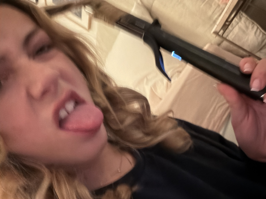 A blurry, close-up selfie captured indoors depicts a light-skinned individual with wavy blonde hair, winking with the right eye and extending their tongue. The subject is wearing a dark, possibly navy blue, long-sleeved top and a delicate necklace with a small, circular pendant. Their right hand, adorned with nails manicured in a French tip style, holds a black hair curling iron or wand. The tool displays a prominent blue illuminated button, indicating it is powered on or heating. The background is softly lit and out of focus, revealing light-colored bedding, likely on a bed, and a partially visible framed object on a surface in the upper right. The scene is located in a room in the city of Paris, France.