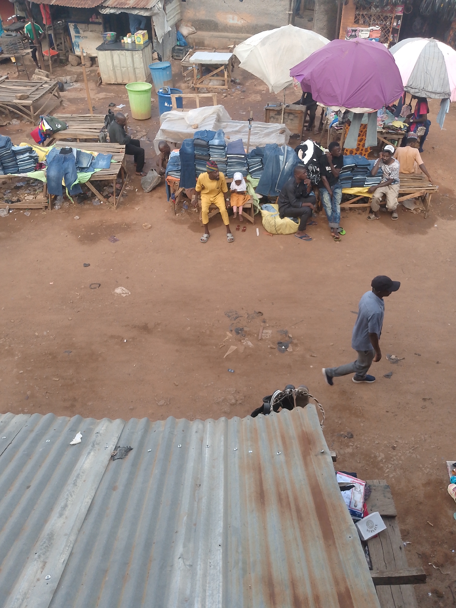 This image depicts a bustling marketplace in Bukuru, Nigeria. The scene is viewed from an elevated perspective, looking down upon a dirt ground area filled with vendors and shoppers. Several stalls display stacks of denim jeans, arranged on wooden benches. Underneath colorful umbrellas (white and purple), groups of men are sitting on the benches, presumably either vendors or potential customers. One man wears a bright yellow outfit while a young child in a white outfit sits beside him. In the foreground, a man wearing a blue shirt and baseball cap is walking. At the bottom of the image is a corrugated metal roof. The overall environment appears busy, with evidence of both commercial activity and pedestrian traffic. There is also a green and blue trash bin near one of the stalls.
