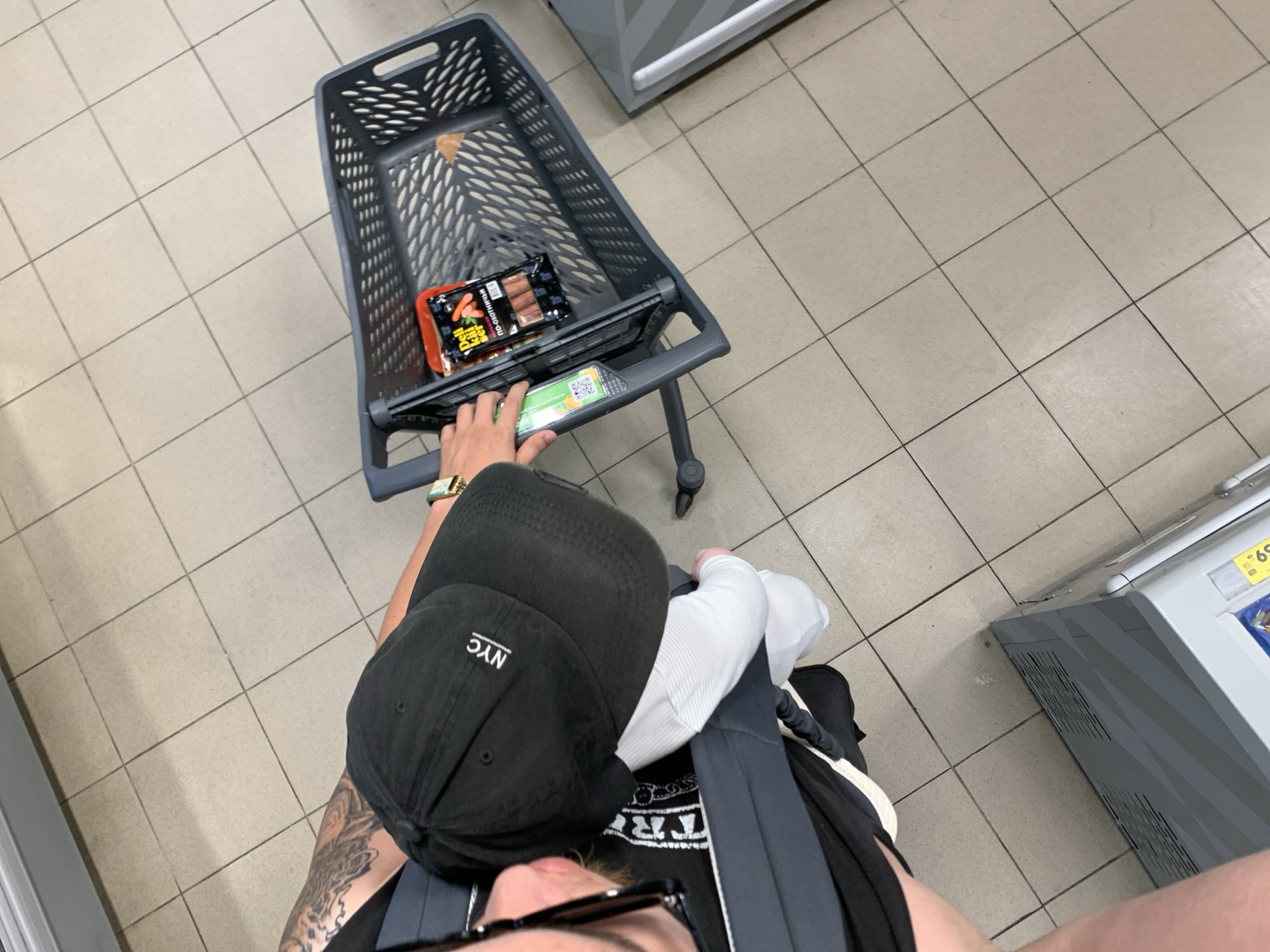 The image shows a person wearing a black baseball cap with "NYC" embroidered on it, pushing a shopping cart in what appears to be a supermarket. The shopping cart contains a package of sausages. The person is also carrying a child in a baby carrier. The floor is tiled. The image does not provide any information about the city of Stavropol, Russia.