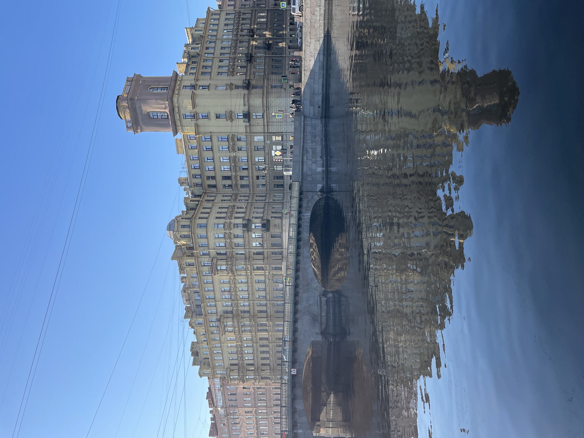The image displays a daytime scene in Saint Petersburg, Russia, centered on the House with Towers (Dom s bashnyami) at 33 Kamennoostrovsky Avenue, overlooking the Karpovka River. The multi-story building, constructed in a light beige or yellow hue, features numerous windows and decorative elements. A prominent multi-story tower with a square base, a central section containing arched windows, and a smaller, rounded upper observation level rises from the building's right side. To the left, another multi-story building with a reddish-pink facade is partially visible.
The Kamennoostrovsky Bridge, constructed of light gray stone with multiple arches, spans the Karpovka River in the mid-ground. A dark metal railing runs along the bridge's top edge, and a white numerical marker displaying "3.4" is affixed to the bridge structure. A street extends across the bridge and along the river embankment, where several pedestrians are visible standing or walking. A few vehicles, including a white SUV, are parked or in transit on the street. Traffic signs, including pedestrian crossing markers, are positioned along the street. Overhead utility wires traverse the clear blue sky. The calm, dark surface of the Karpovka River occupies the lower half of the image, reflecting the buildings, bridge, and sky with slight distortions. The scene is illuminated by bright natural light, casting subtle shadows.