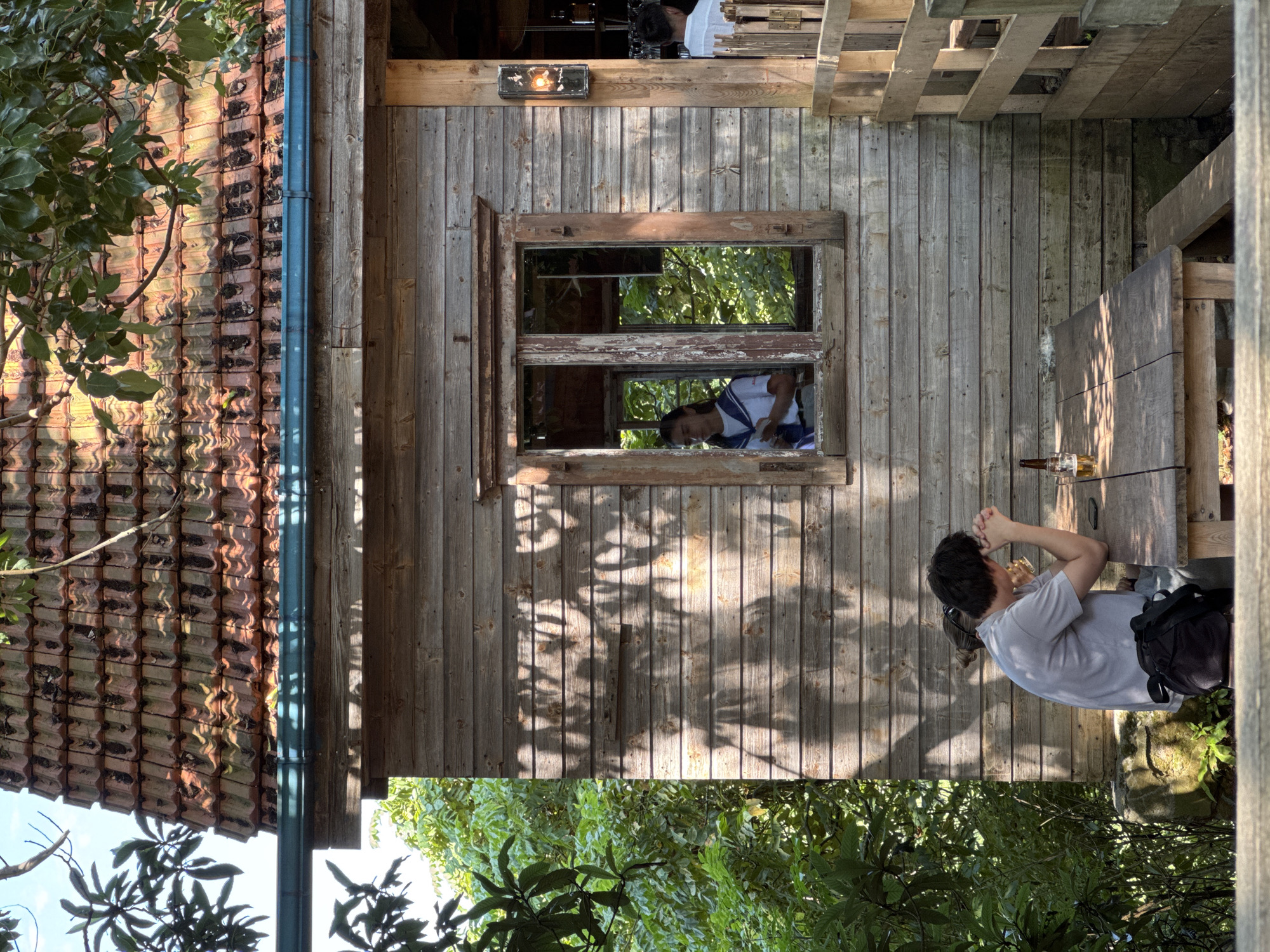 In Buknari, Georgia, a rustic wooden structure with a tiled roof is depicted. In the foreground, a man is seated at a wooden table, his hands clasped in front of him, appearing contemplative. A bottle of beer and a glass with beer are visible on the table. To the left of the man, lush green foliage suggests an outdoor, natural setting. The building features a window through which a woman can be seen inside, holding a phone. The overall scene evokes a relaxed, casual atmosphere, possibly a cafe or a resting spot in a natural environment.